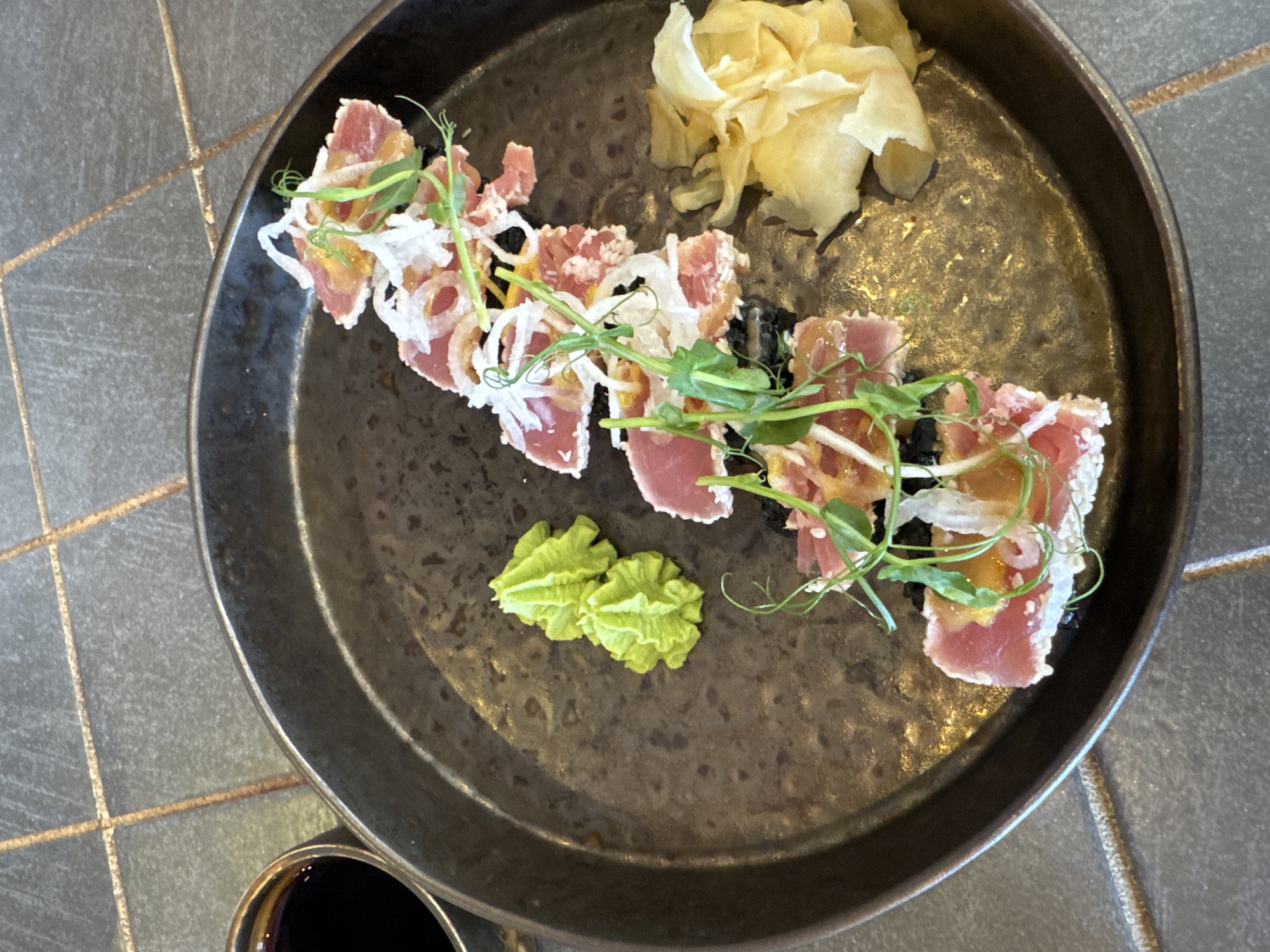 The image shows a plate of seared tuna sashimi, likely served in a restaurant in an unknown city. The dish is arranged on a dark, textured plate. The sashimi is garnished with white onion rings, pea shoots, and a small mound of wasabi. Pickled ginger is also present on the plate. A small container of soy sauce is visible in the corner of the image. The plate is placed on a tile-covered surface.