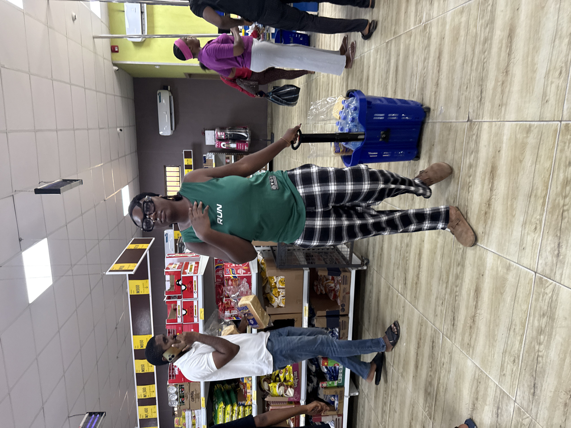 A view inside a grocery store in Ado, Nigeria. A person with glasses wearing a green tank top with the word "RUN" on it is seen pulling a blue shopping cart filled with bottled water. They are wearing black and white plaid pants and brown shoes. A man in a white shirt and jeans is seen standing near shelves of groceries and talking on a mobile phone. Another person wearing a purple shirt is seen behind the shopping cart. Price tags are visible on the shelves.