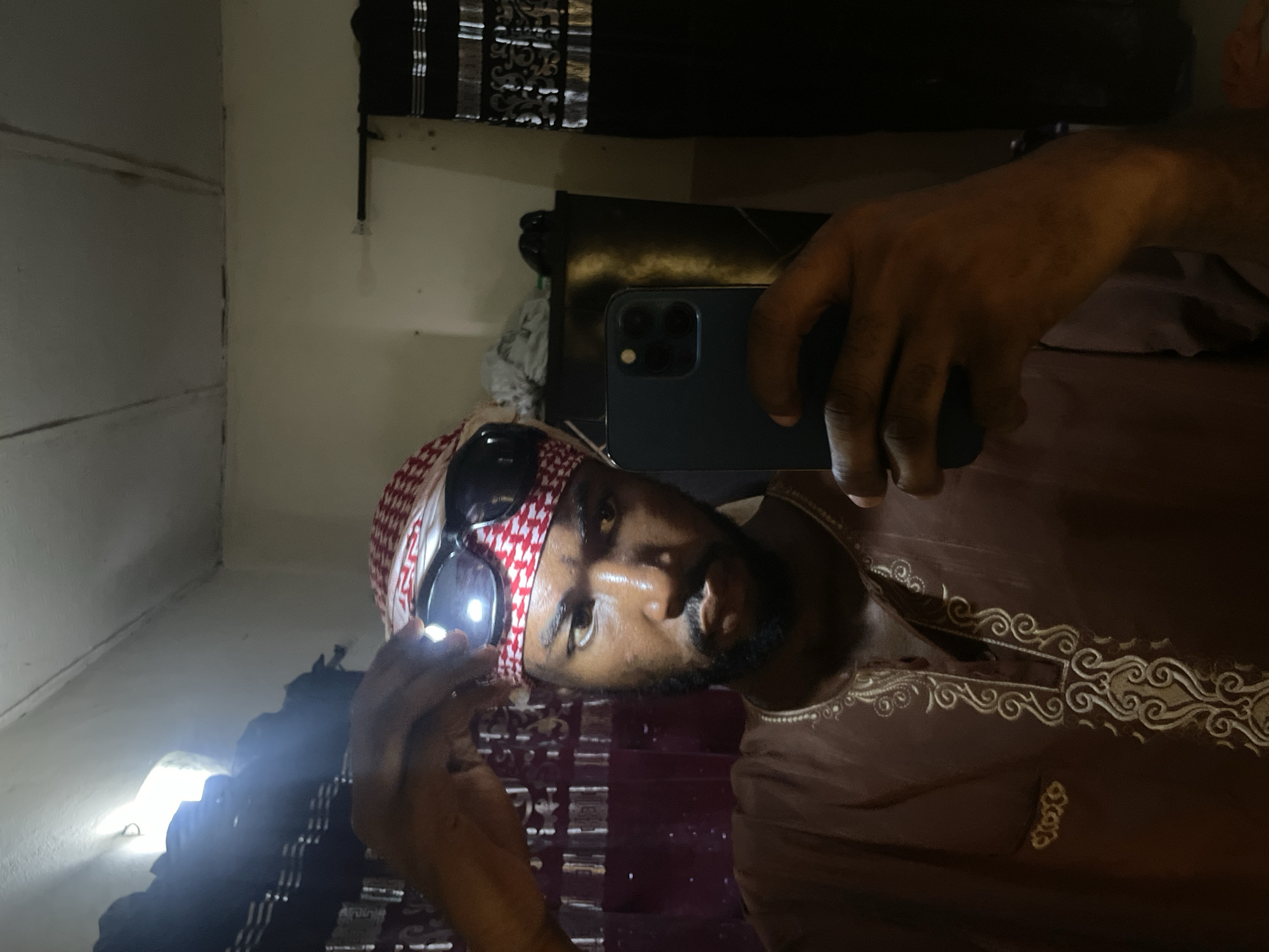A man wearing sunglasses and a red and white keffiyeh is taking a selfie in a room. He's holding a smartphone in his right hand. He's wearing a brown robe with ornate gold embroidery around the neck. The room appears to be dimly lit and the setting appears to be a bedroom. Lokoja, Nigeria is not discernable from the image.