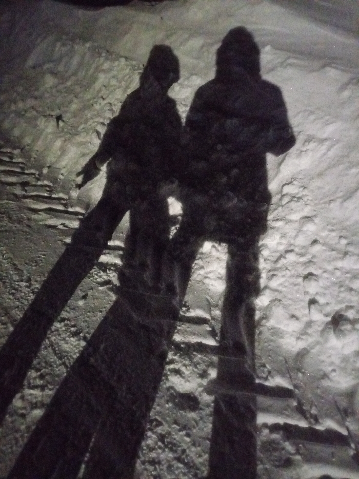 The image depicts a nighttime outdoor scene in the city of Старый Шагирт, Russia. Two human figures are visible as dark silhouettes against a snow-covered environment, illuminated from behind by an unseen light source that casts long, distinct shadows towards the viewer. The figure on the left is smaller, with its right arm slightly extended forward. The figure on the right is taller and stands upright. Both figures appear to be wearing hooded garments. The ground is uneven, entirely covered in snow of varying depths and textures, with visible footprints or depressions in certain areas. No other distinct objects or specific actions are discernible.