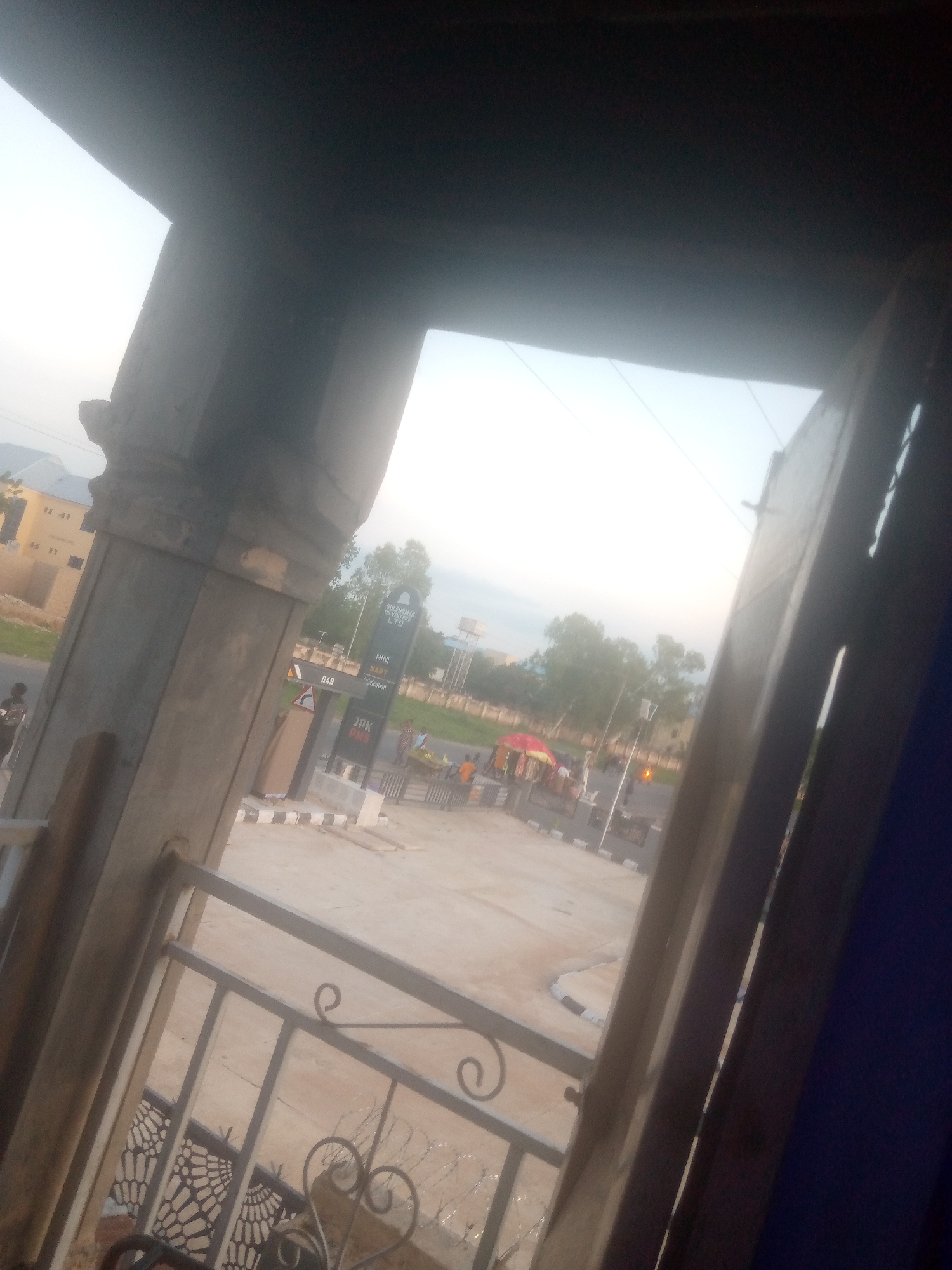 The image shows a view from an interior space looking out onto a street in Jalingo, Nigeria. In the foreground, a metal railing with decorative scrolls is visible. To the left, a large concrete pillar with ornate carvings is present. The background reveals a street scene with a gas station, identified by a sign for "SULTANAH BENENINE LTD" with smaller signs indicating "MINI MART" and "Lubrication." A "GAS" sign is also prominent. Several people are scattered around the gas station, some near a red and yellow umbrella. Buildings and trees are visible further in the background, with a water tank on a pole in the distance. The sky is overcast and bright, suggesting either early morning or late afternoon light. The overall scene appears to be a typical urban streetscape in Nigeria.