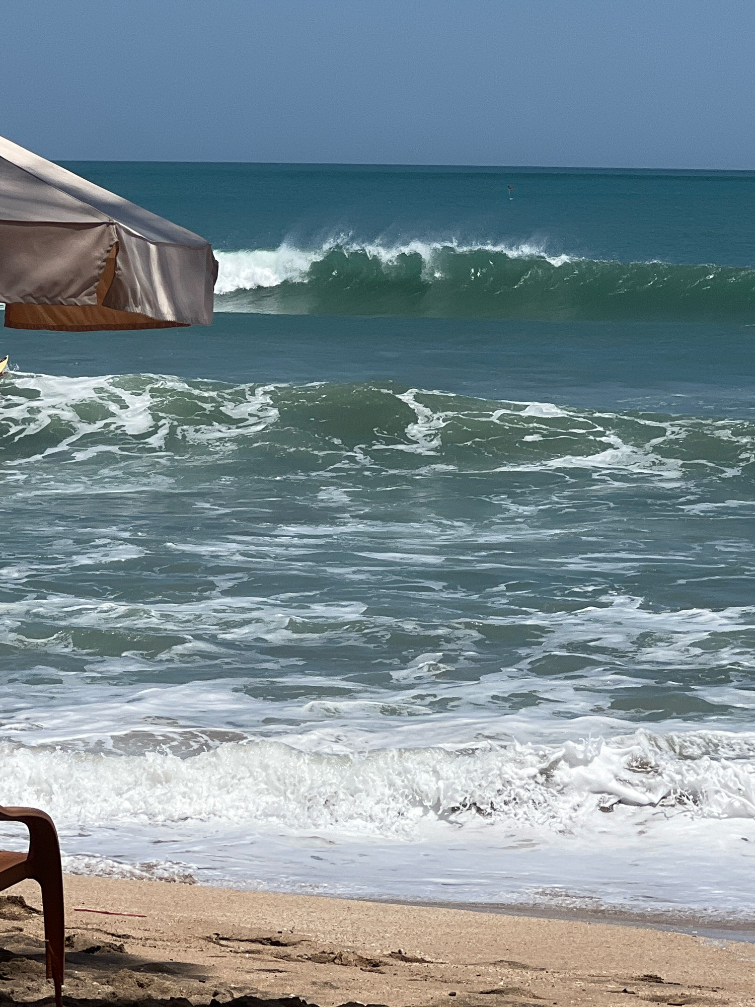 A wide shot of Kuta Beach in Bali, Indonesia, shows a choppy ocean with breaking waves. In the foreground, a portion of a beach umbrella and a brown plastic chair are visible. The umbrella is made of a light-colored fabric with a darker trim. The ocean water is a deep blue-green, with white foam from the waves. Further out, a larger wave is breaking, with spray visible in the air. The sky is a clear, light blue. In the distance, a small red flag is visible on a pole in the ocean. The sand on the beach is light brown and appears wet in places.