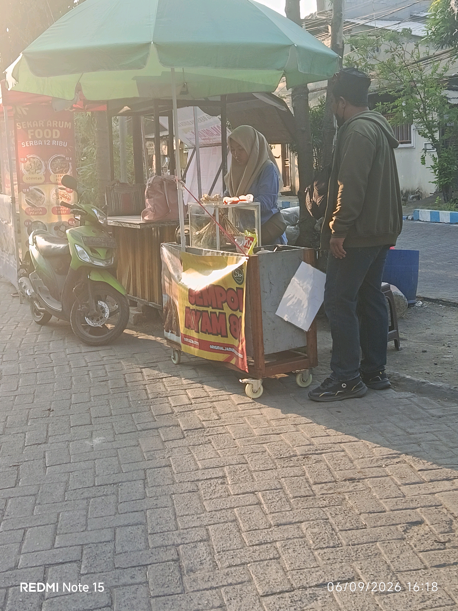 In the city of Keboansikep, Indonesia, at 4:18 PM on September 6, 2026, a street food vendor, a woman wearing a light brown hijab and a blue shirt, stands behind a cart selling food. The cart is adorned with a large yellow banner featuring red text that reads "CEMPOL AYAM 8 RIBU". To the right of the vendor, a man wearing a dark green hoodie and dark pants stands facing her, appearing to be a customer. A green scooter is parked to the left of the food cart. The scene takes place on a paved sidewalk with a sunlit ambiance, casting shadows. Behind the vendor and cart, a green umbrella provides shade. The overall environment suggests a typical outdoor market or roadside stall setting.