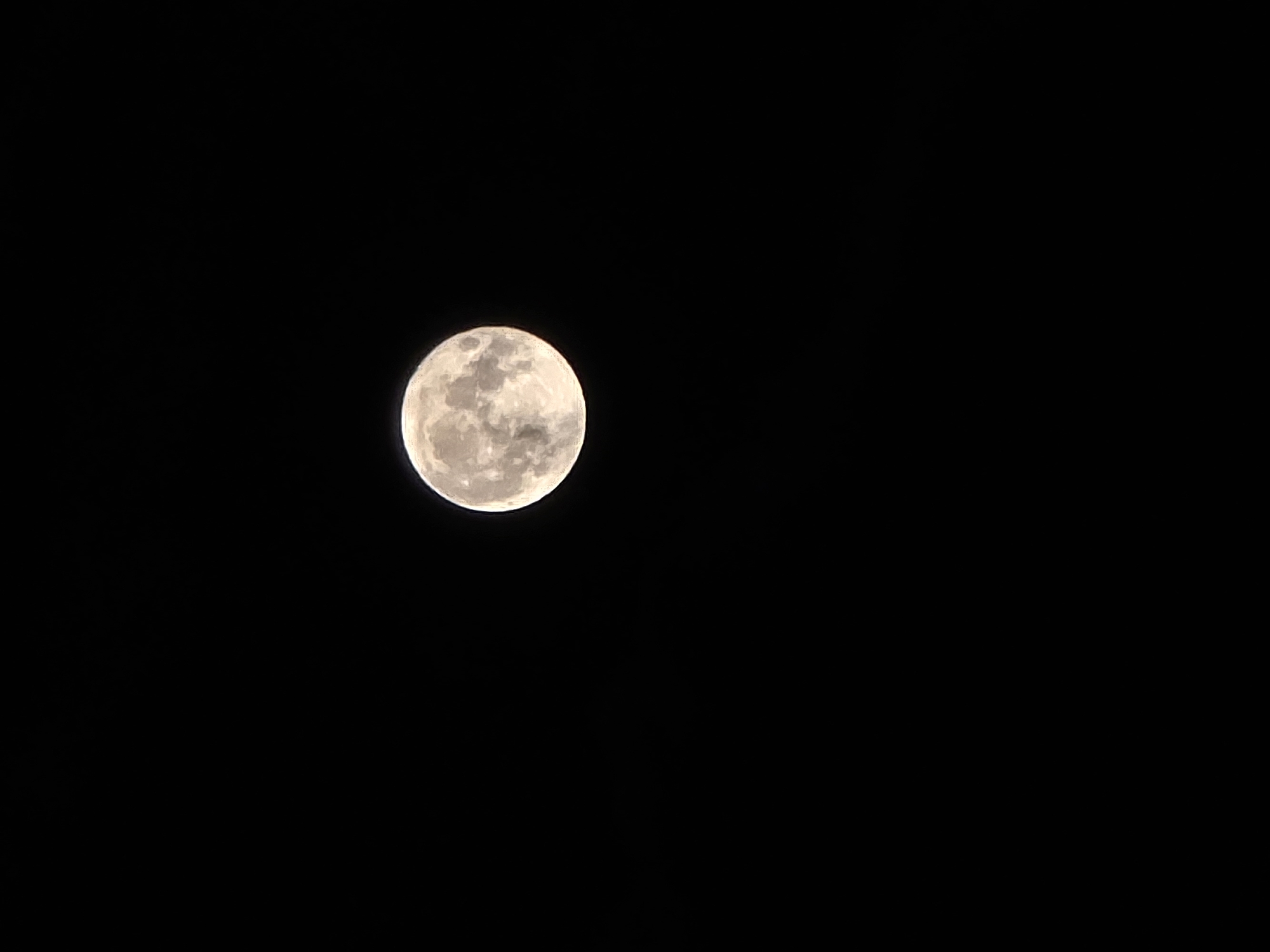 The image shows a full moon against a black night sky. The moon is centered and slightly to the left. It displays visible craters and surface texture. The background is completely dark. No specific location or city can be determined from this image.