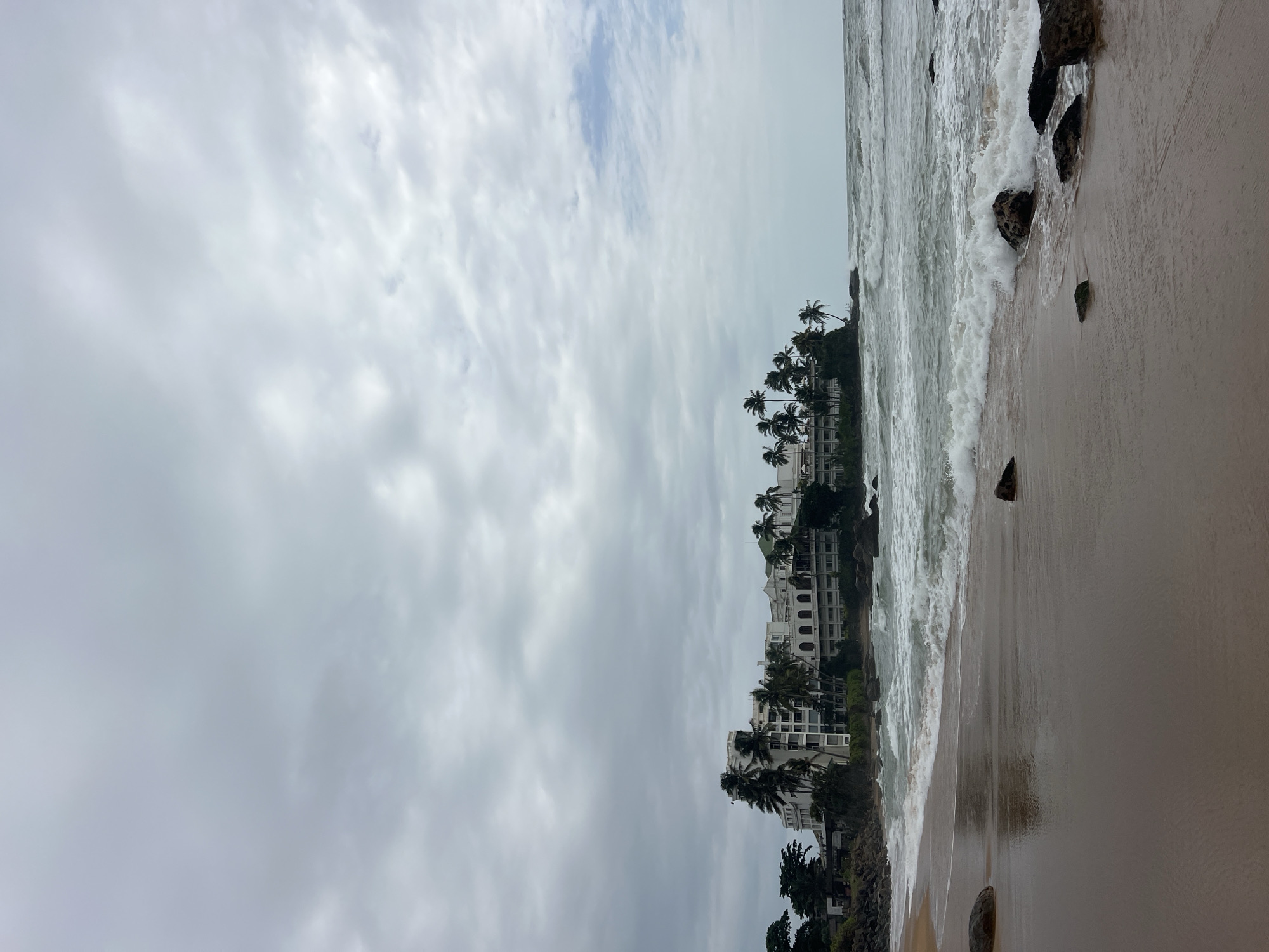 A wide shot captures a cloudy day on the beach in Dehiwala, Sri Lanka. In the distance, a white hotel with multiple levels and arched windows sits on a rocky outcrop surrounded by palm trees. The ocean in front of the hotel has moderate waves crashing onto the shore. The beach in the foreground is wet sand with scattered rocks, some of which are partially submerged by the incoming tide. The sky is filled with heavy gray clouds, creating an overcast atmosphere.