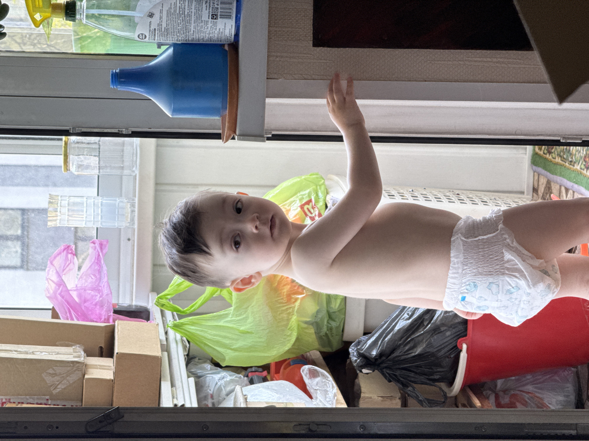 A baby wearing a diaper lies on a variety of objects, including boxes, plastic bags, and a red bucket. The location is Izhevsk, Russia. Other objects present include cleaning supplies, glass jars, and a black trash bag. The baby's left arm is raised. The background consists of a window and a partially visible rug.