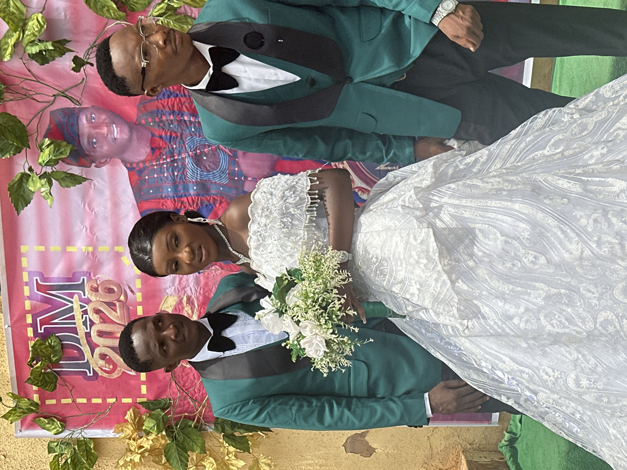 The image shows a bride and two grooms posing in front of a banner. The banner is pink and has the letters "DM 2020" on it. The bride is wearing a white wedding dress and holding a bouquet of white flowers. The two grooms are wearing green suits with black bowties. The location is Unknown, Unknown. There are artificial flowers and green leaves used as decoration. One of the grooms is wearing glasses and a wristwatch. There is a picture of a person behind the wedding couple on the banner.