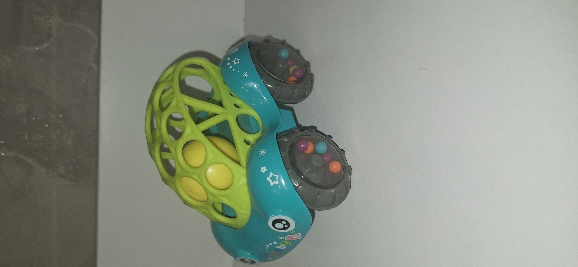 A close-up, eye-level shot shows a turquoise and lime green baby rattle shaped like a car, positioned on a plain white surface against a gray, mottled background. The car rattle features large, grey wheels that contain colorful beads, including orange, purple, and light blue. The body of the car is turquoise with black eyes and a pink and white sticker with the word "baby" printed on it. A lime green, open-cage structure sits atop the car's body, enclosing a bright yellow ball. The overall lighting is neutral, highlighting the toy without casting harsh shadows. There are no people or discernible elements of the city of Bila Tserkva, Ukraine present in this image.