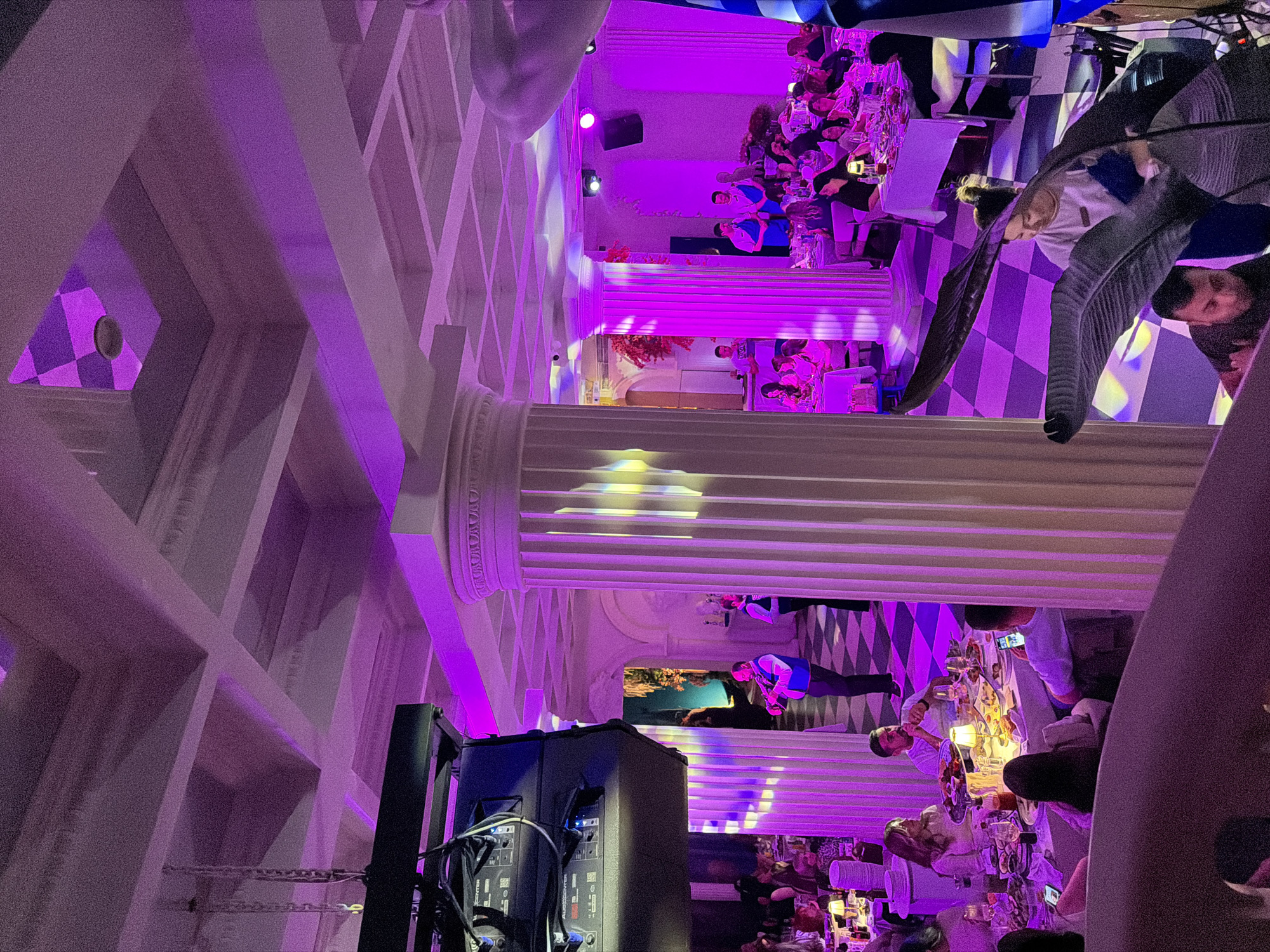 The image depicts the interior of a restaurant, likely in Yerevan, Armenia, characterized by elaborate, Greek-inspired architectural elements such as columns, checkerboard-patterned walls, and a white coffered ceiling with triangular recesses. The restaurant features a crowded dining area with multiple tables occupied by people engaged in dining and socializing. The lighting is primarily purple, creating a vibrant ambiance. A speaker or sound system is visible in the foreground, suggesting the presence of music or entertainment. The image is rotated 90 degrees.