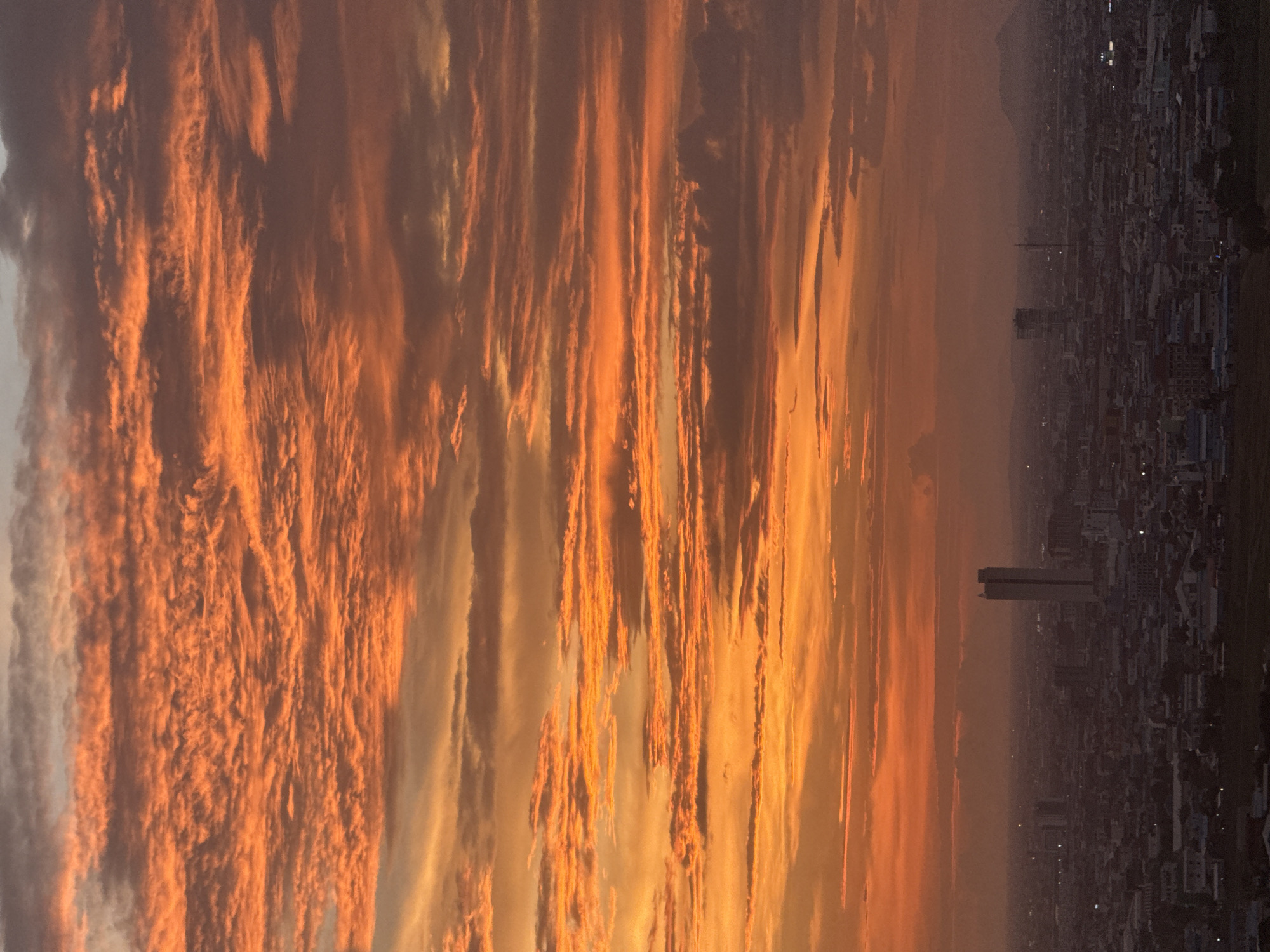 The image depicts a panoramic view of the city of Khan Mean Chey, Cambodia, at sunset. The sky is dominated by vibrant orange and red hues, with numerous elongated clouds creating a textured pattern across the horizon. In the foreground, the city sprawls across the landscape, a dense collection of buildings and structures. A prominent tall, rectangular building stands out in the mid-ground, with other smaller buildings and the outlines of distant hills visible further back. The lighting suggests it is late evening or early morning, with the city lights beginning to twinkle. There are no people or animals visible in the frame.
