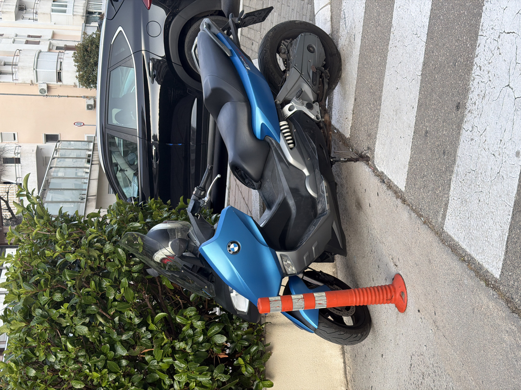 A blue BMW scooter is parked partially on a pedestrian crossing. The scooter is a maxi-scooter style, with a large fairing and a comfortable seating position. It is blue and grey in color, with a black seat. The front of the scooter has a BMW logo. A red and white striped bollard is in front of the scooter, on the pedestrian crossing. Behind the scooter, a black car is parked. To the left of the scooter is a dense green bush. In the background, there are buildings that appear to be apartments or hotels, suggesting an urban or tourist setting. The scene appears to be outdoors, with natural light. The time of day is unclear, but the lighting suggests it is daytime. The location appears to be Budva, Montenegro, given the buildings and the style of the street. There are no people visible in the image. The scooter is the main subject of the image, and it is parked in a way that obstructs the pedestrian crossing.