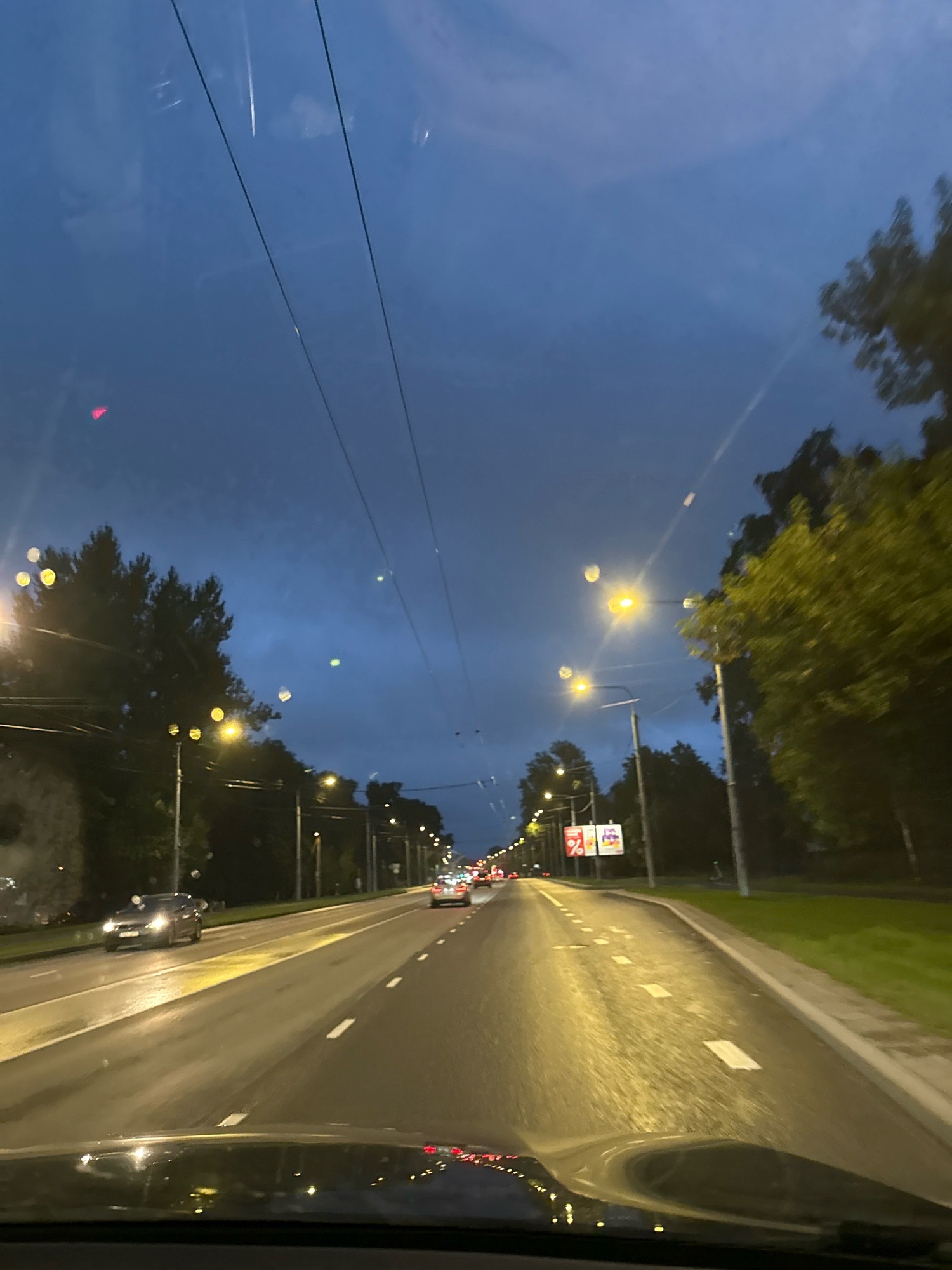 The image depicts a view from a vehicle traveling on a multi-lane road at dusk or early night, likely in Tallinn, Estonia, given the context provided. The sky is a deep blue, with some clouds and power lines visible. Streetlights illuminate the road, which appears wet, reflecting the lights. Trees line the sides of the road. Several cars are visible on the road, some in motion and some parked. There are buildings and signs visible in the distance. The foreground shows the dashboard of the vehicle, also reflecting the streetlights and the road ahead. The overall impression is a typical urban driving scene at twilight.