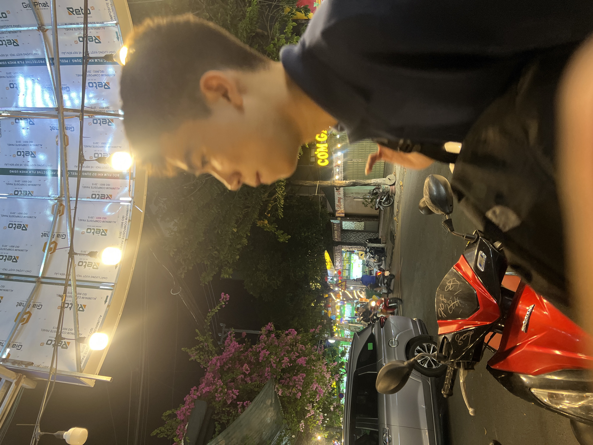 In a nighttime urban scene in Da Nang, Vietnam, a young man is captured in profile, looking to his right. He wears a dark blue collared shirt. The background reveals a street with parked and moving motorcycles, a silver car, and several shops with illuminated signage. Pink bougainvillea flowers cascade from overhead, and exposed light bulbs hang from a structure above. One motorcycle in the foreground is red and black with visible markings on its front. The setting suggests a lively street at night, possibly near a commercial area.