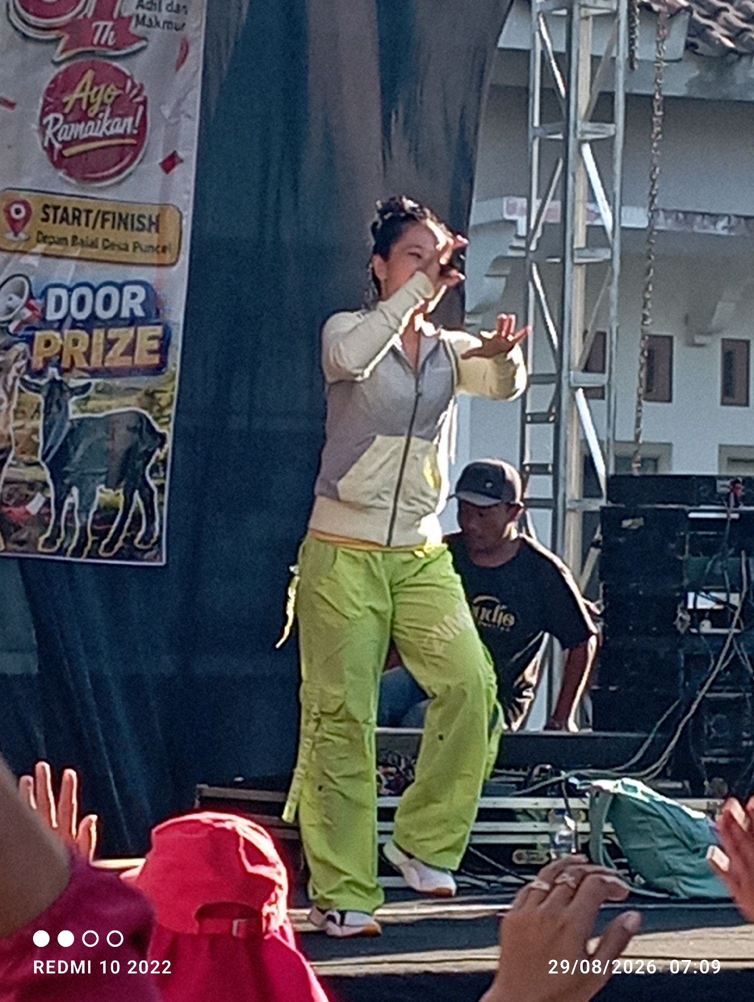 The image captures an outdoor event, likely in Dukuhseti, Indonesia, with a stage set up. In the foreground, several hands are visible, some raised. A person in a red hat is partially seen, and what appears to be a green backpack lies on the stage.
On the stage, a woman with dark hair tied back is performing, wearing a light-colored jacket with grey accents and bright green cargo pants. She is facing left and gesturing with her hands. To her right, a man wearing a dark t-shirt and a baseball cap is looking down. Behind them, a stage structure and audio equipment are visible, along with a banner that includes text in Indonesian, such as "START/FINISH" and "DOOR PRIZE", and an image of a goat. The environment appears to be sunny, suggesting daytime.