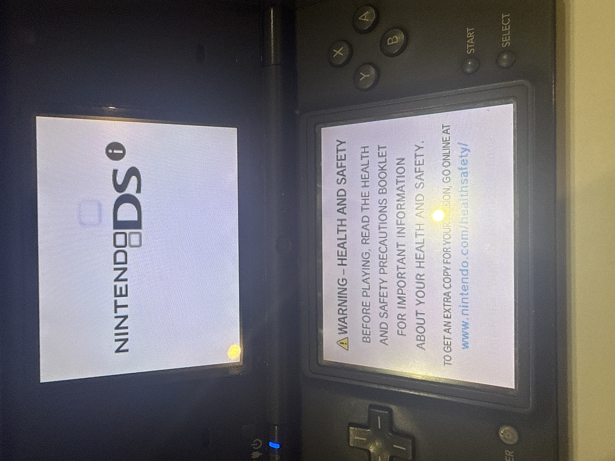 An overhead shot shows a black Nintendo DS console displaying a health and safety warning. The top screen shows the Nintendo DS logo with the text "WARNING - HEALTH AND SAFETY", followed by instructions and a website address. The bottom screen has directional buttons, a power button, a blue light, and other controls. The console is on a light-colored surface. The image is in focus and well-lit, with no identifiable people or specific locations within the city of Burgas, Bulgaria.