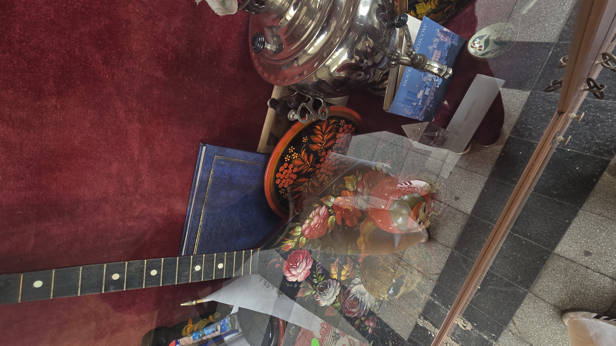 The image shows a glass display case containing an assortment of objects. Among them are a balalaika with a floral design, a silver samovar, a black and red lacquered tray, a blue book, a postcard with an image of Moscow, and a small ceramic bowl with a floral pattern. The display case is placed on a floor with black and grey tiles. A portion of a shoe is visible outside the display case, suggesting that it is located in a public space. The reflection in the glass adds additional layers of visual information. Without further context, it is impossible to confirm the precise location in Stara Zagora, Bulgaria.