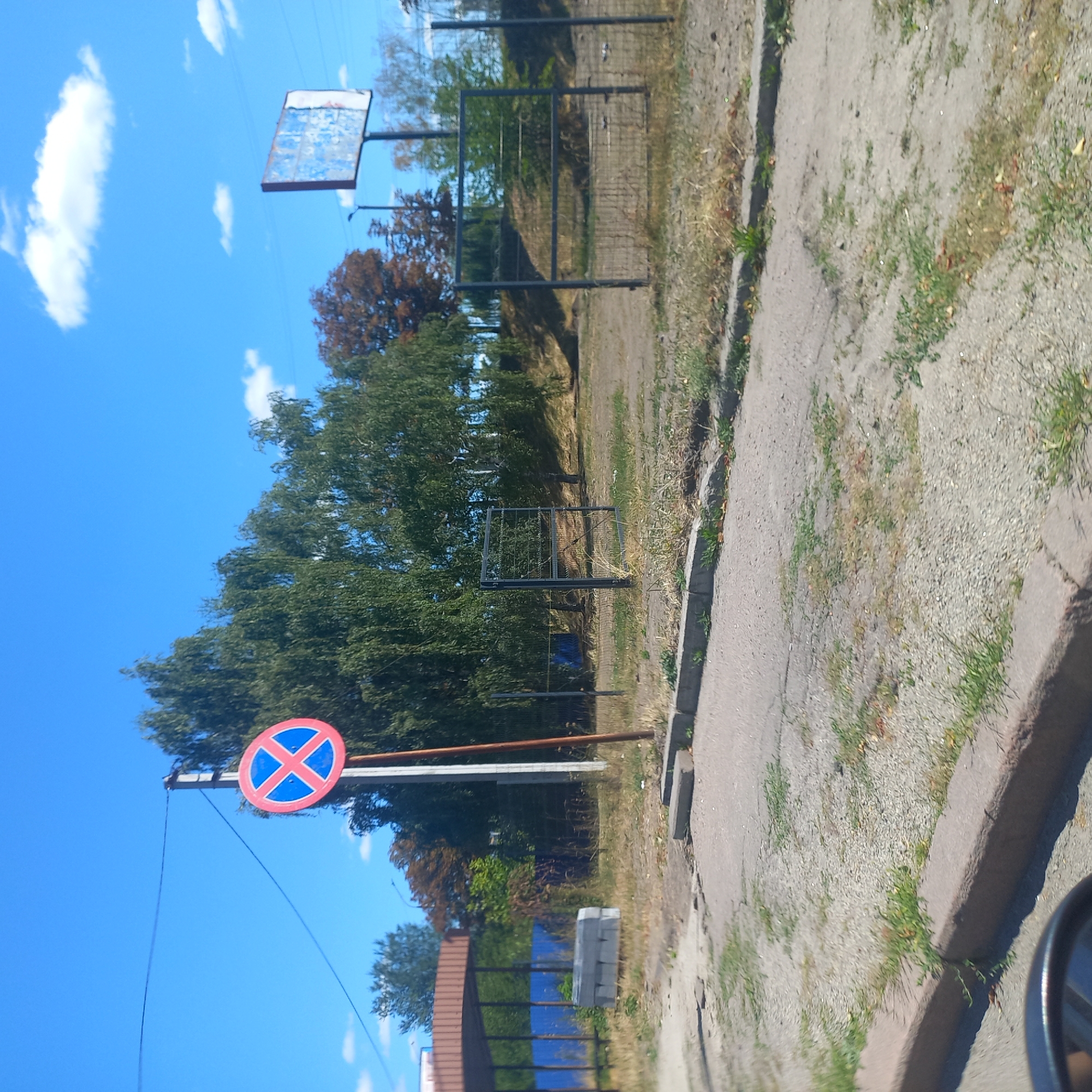 The image depicts a dry, outdoor scene under a clear blue sky with scattered white clouds. In the foreground, a broken asphalt surface is visible with patches of dry grass and weeds growing through cracks. A concrete curb runs along the edge of the asphalt.
Dominating the left side of the image is a circular "No Parking" sign with a red border and a blue interior featuring a red "X". The sign is mounted on a slender wooden pole, which is next to a utilitarian concrete utility pole. Behind the sign, a section of a brown-roofed shelter with a blue fence is visible, along with stacks of grey concrete blocks.
To the right of the "No Parking" sign, a black metal gate stands open, leading into a grassy area with a gentle slope. Beyond this, more trees are visible, including some with autumn foliage, and a large, weathered billboard with blue and white markings is mounted on a dark pole. The overall environment suggests a somewhat neglected urban or industrial fringe area.
The specific location in Kropyvnytskyi, Ukraine, is not discernible from the visual elements present in the image itself.