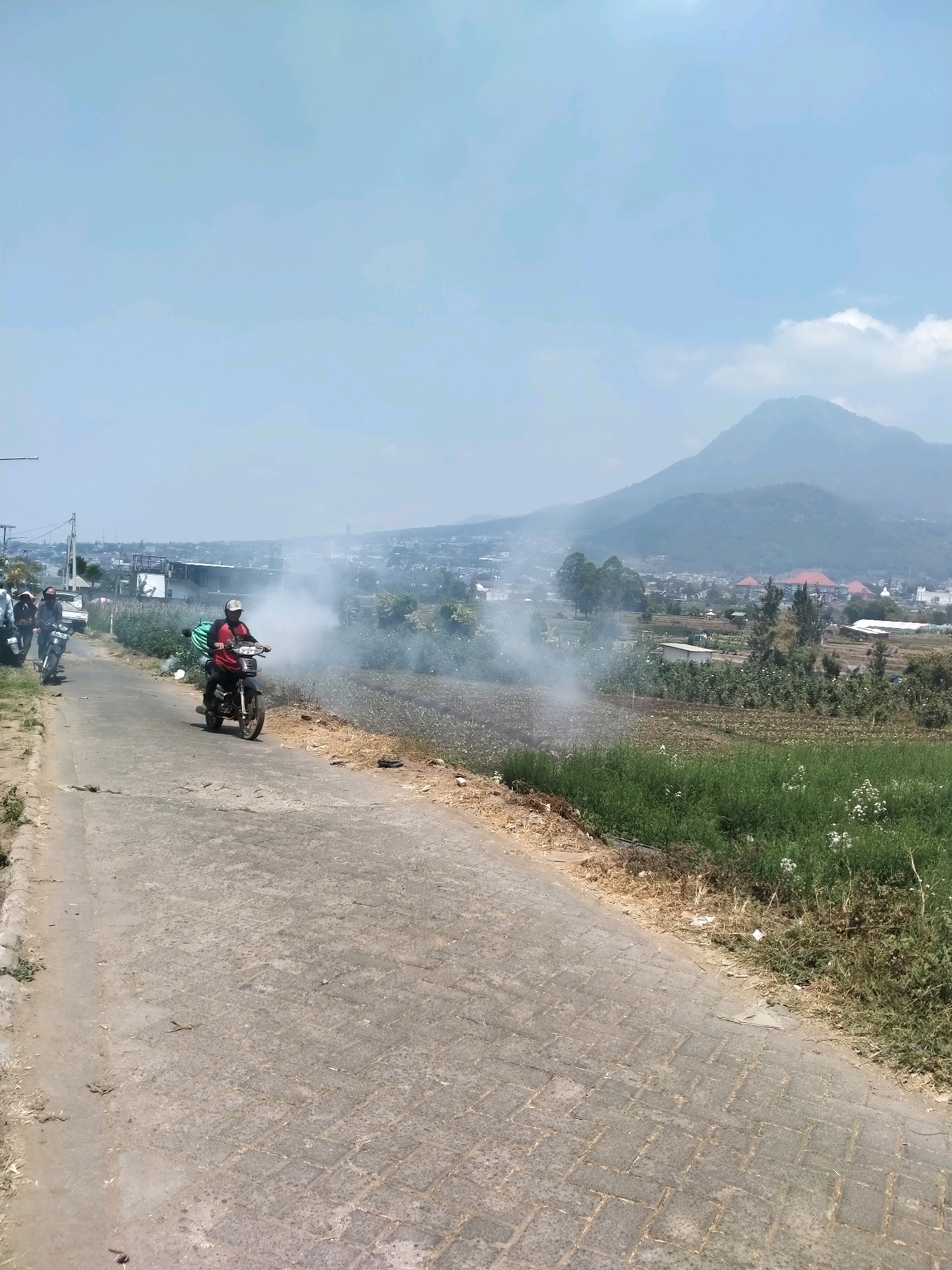 A motorcyclist wearing a red shirt and black pants is seen riding a motorcycle on a paved road. They are surrounded by a cloud of white smoke. The road curves to the right, leading towards a field with crops and vegetation. In the background, a town or village is visible, with buildings and houses nestled at the base of a large, misty mountain. The sky is clear and blue with some scattered clouds. The overall scene depicts a rural or semi-urban environment with agricultural activity and a prominent natural landscape.