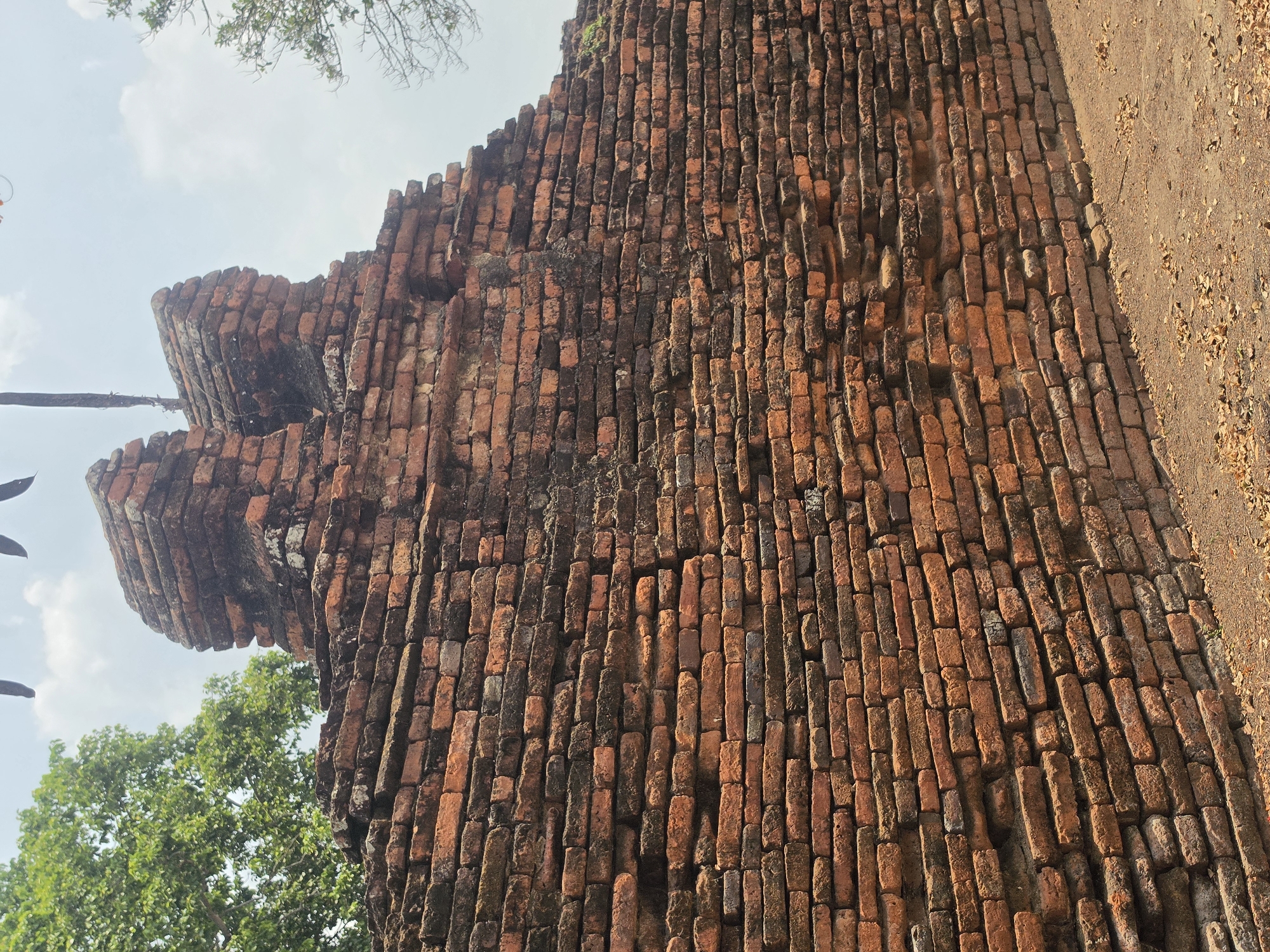 An upward-angled view captures a large, ancient brick wall structure, likely part of a historical ruin in Phra Nakhon Si Ayutthaya City Municipality, Thailand. The wall dominates the frame, showcasing layers of weathered, reddish-brown bricks. Many bricks exhibit discoloration, dark stains, and signs of erosion, indicating significant age.
On the left side of the image, the brick structure tapers upwards, featuring distinct tiered or corbelled sections that suggest an architectural detail of a tower or building edge. Green foliage from trees is visible on the far left and at the bottom left, partially obscuring the base of the structure and framing the top against a bright, clear sky with soft white clouds. The right side of the main wall extends into the distance, meeting a patch of sandy, earthy ground covered with scattered dry leaves.
No human activity is depicted. The scene suggests a bright, clear daytime. There is no visible text. The overall impression is one of ancient history and architectural resilience against time.
