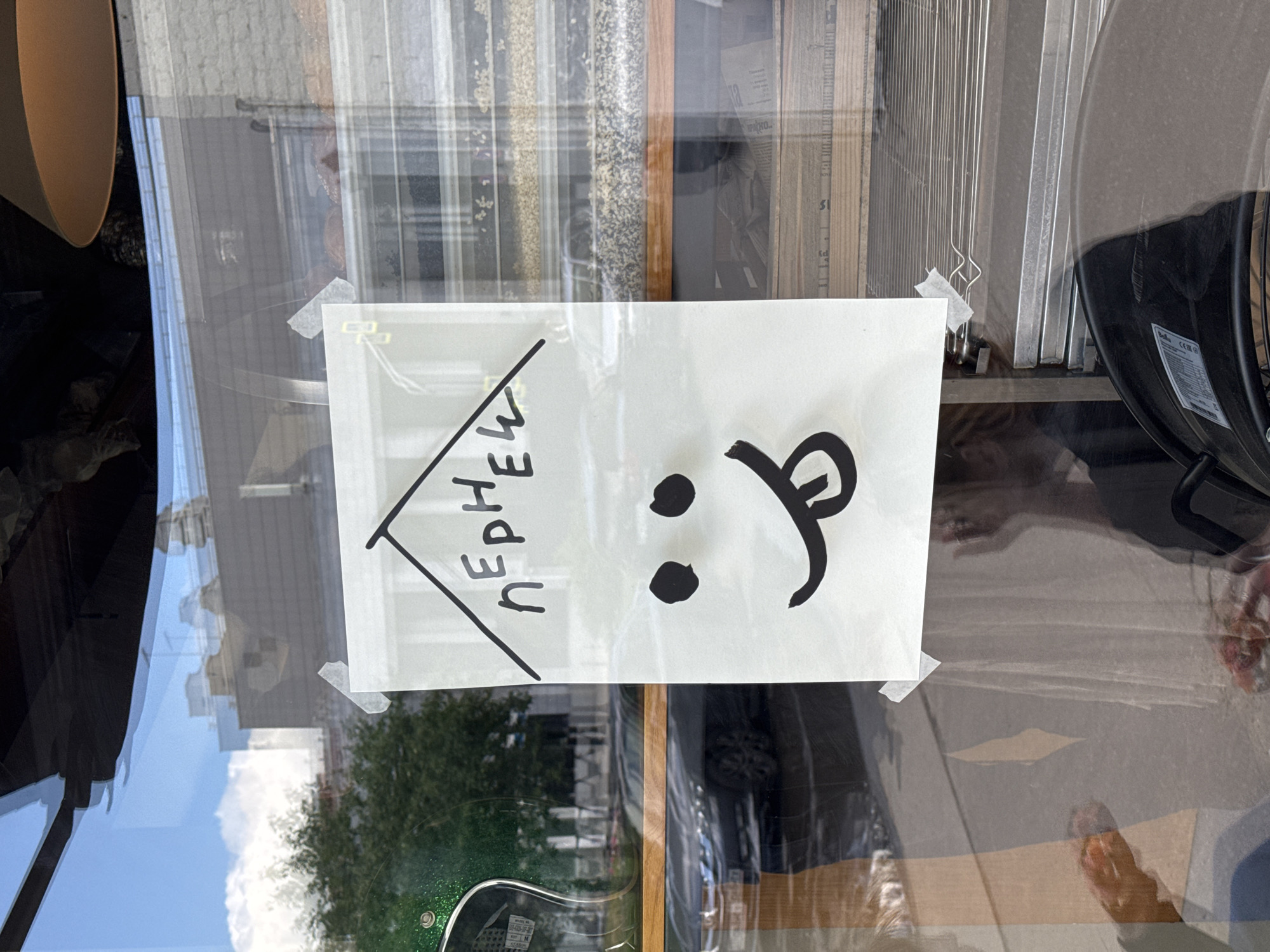 The image shows a hand-drawn smiley face on a piece of paper taped to a glass window. The smiley face has two black dots for eyes and a wide smile with its tongue sticking out. Above the smiley face, also drawn on the paper, are Cyrillic letters that appear to form the word "ПЕРЕЦ" (PERETS), which means "pepper" in Russian. The paper is taped to the window with white tape, and the drawing is done in black marker. Reflections on the window show a street scene with trees, a car, and buildings, suggesting an urban environment. Inside the establishment, shelves with various items, possibly food products, are visible. The overall impression is a quirky and informal sign. The image's metadata or context is not available to specify the exact location within Moscow, Russia, but the visual cues like the Cyrillic script and the architectural style of the buildings in the reflection suggest this region.
