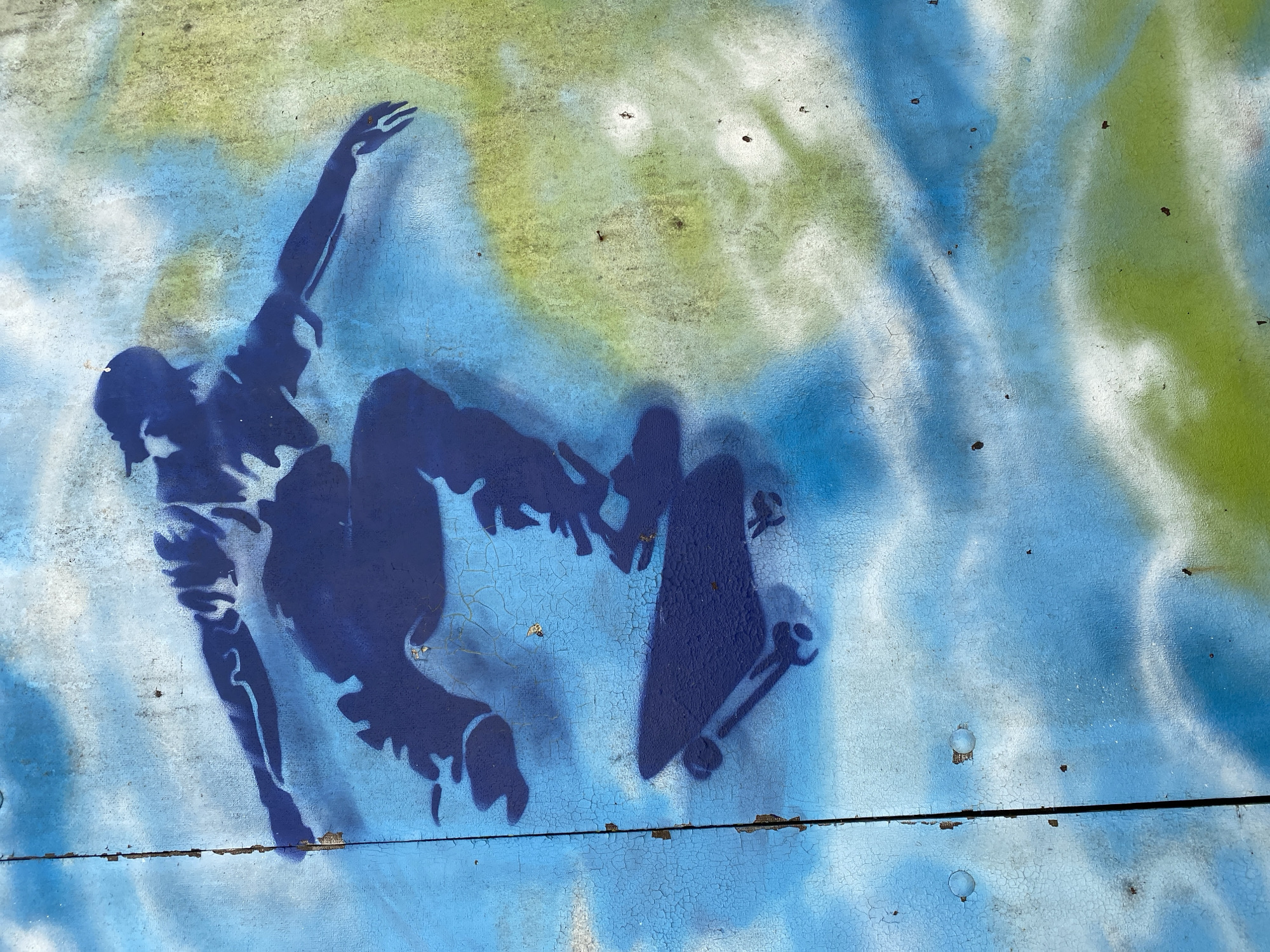 A spray-painted silhouette of a skateboarder in mid-air is depicted on a weathered, blue and green surface. The skateboarder is wearing a baseball cap and appears to be performing a trick. The surface shows signs of age, with peeling paint and visible cracks. There are also some small dark specks scattered across the surface, possibly dirt or rust. The image is a close-up shot, focusing entirely on the artwork and the condition of the surface it is on. The location is identified as Altenkirchen, Germany.