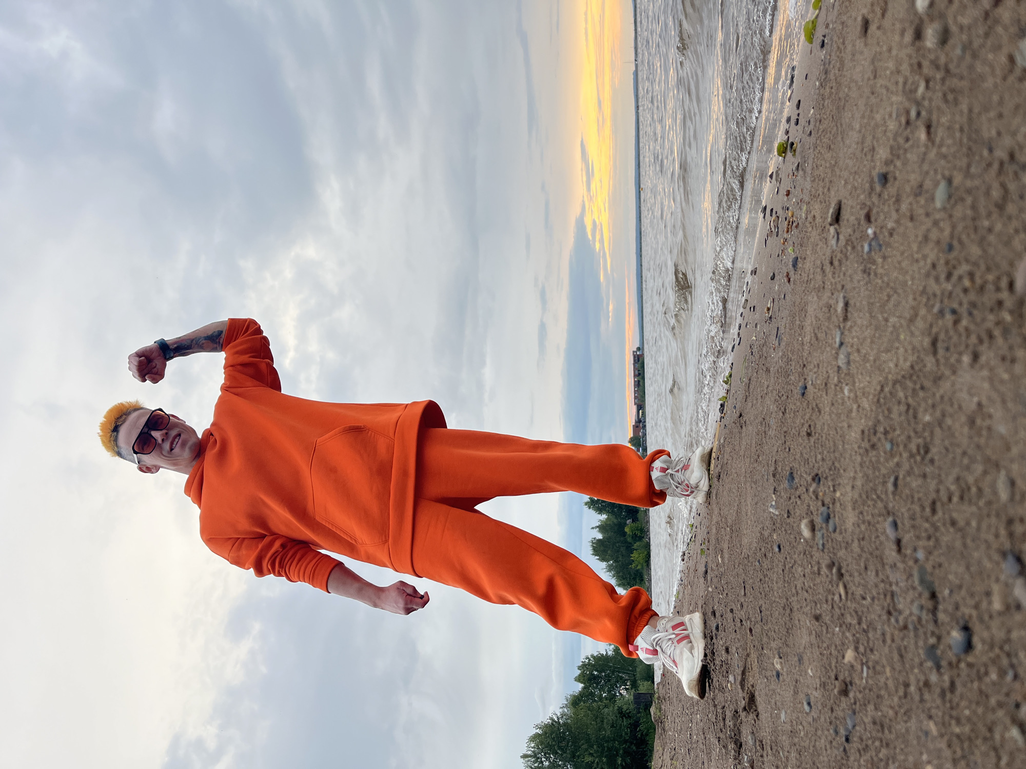 A person with bright orange dyed hair, wearing sunglasses, an orange hoodie, orange sweatpants, and white sneakers with red accents, stands on a sandy beach next to a body of water. Their left arm is raised in a fist, and they are looking towards the camera with a slight smile. The scene is set at sunset, with the sky filled with colorful clouds transitioning from gray to yellow and orange. The water appears calm with small waves lapping at the shore, which is covered in sand and small pebbles. Lush green trees can be seen in the background on the far bank of the water. The image is taken from a low angle, with the subject diagonally positioned in the frame. The overall mood is energetic and casual, possibly indicating a moment of achievement or celebration.