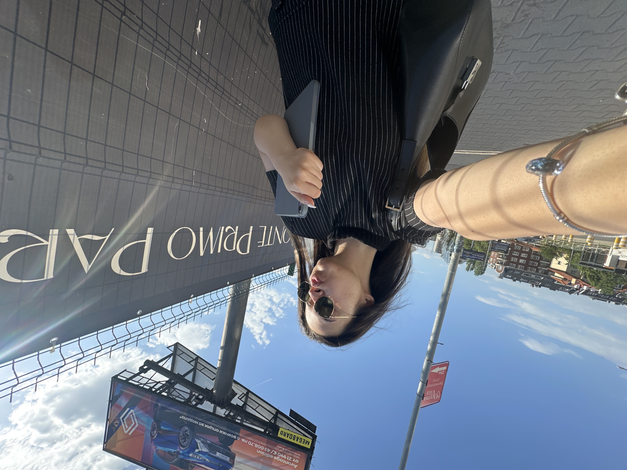The image is an upside-down selfie taken in Sofia, Bulgaria. A woman with long, dark hair, wearing sunglasses and a black and white striped shirt, is holding a gray tablet or phone. She is also carrying a black purse with a buckle strap. Her arm, extended in the selfie, features a silver bracelet.
The background reveals an urban landscape. A large billboard with the words "ONTE PRIMO PAR" is visible above her, as well as a smaller advertising billboard below for a car, featuring text in Bulgarian. A red sign for "Derma Vita Dermatology Clinic" and a directional arrow is also present. The sky is blue with some clouds, and buildings can be seen in the distance. The surroundings suggest a street scene in Sofia.