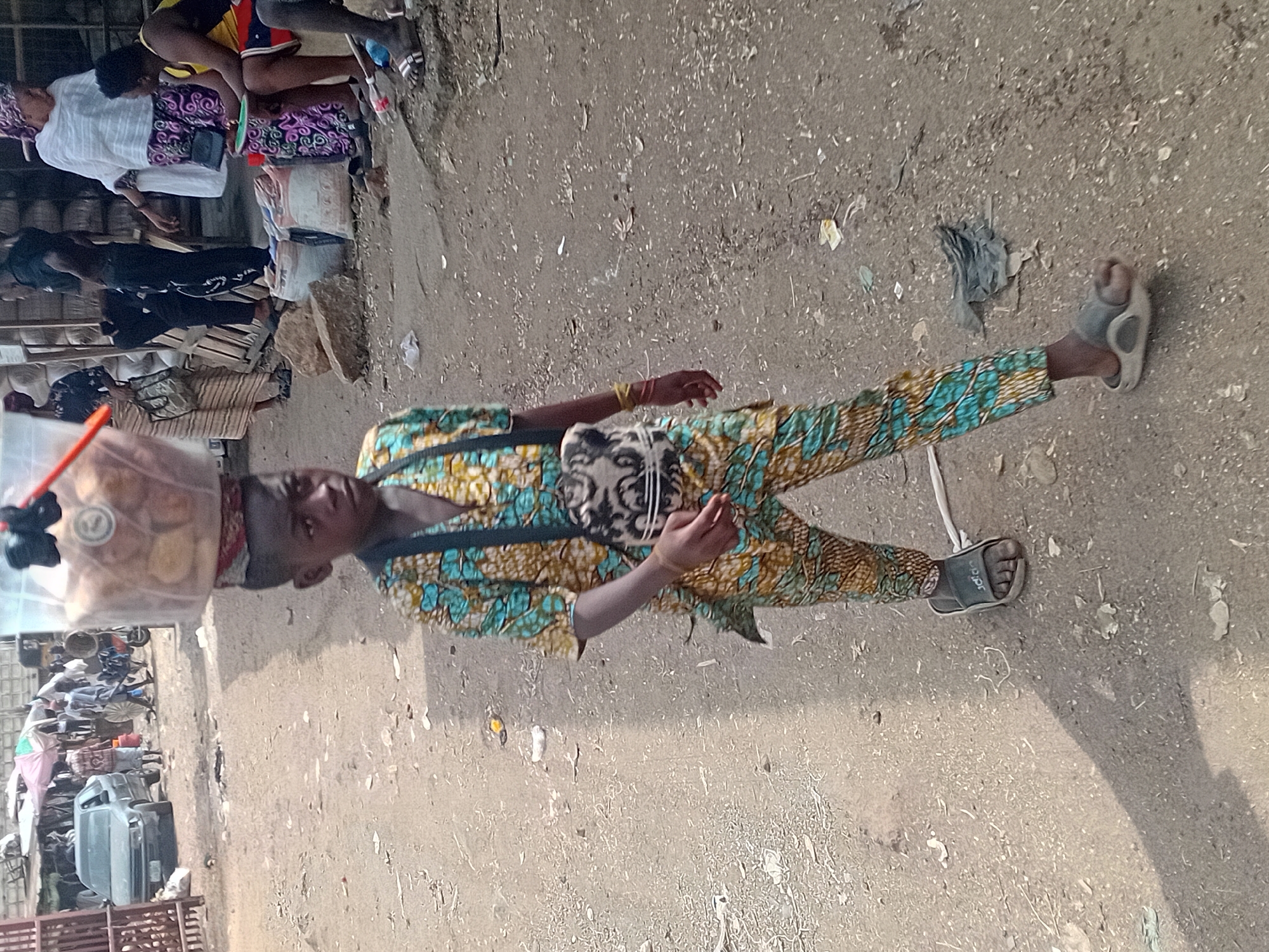 In Lokoja, Nigeria, a young person in a patterned shirt and pants stands on a dirt ground. They are wearing sandals and carrying a bag on their chest. A plastic container filled with food items rests on their head. In the background, a cluttered street scene includes other people, vehicles, and various objects. The setting appears to be an outdoor market area.