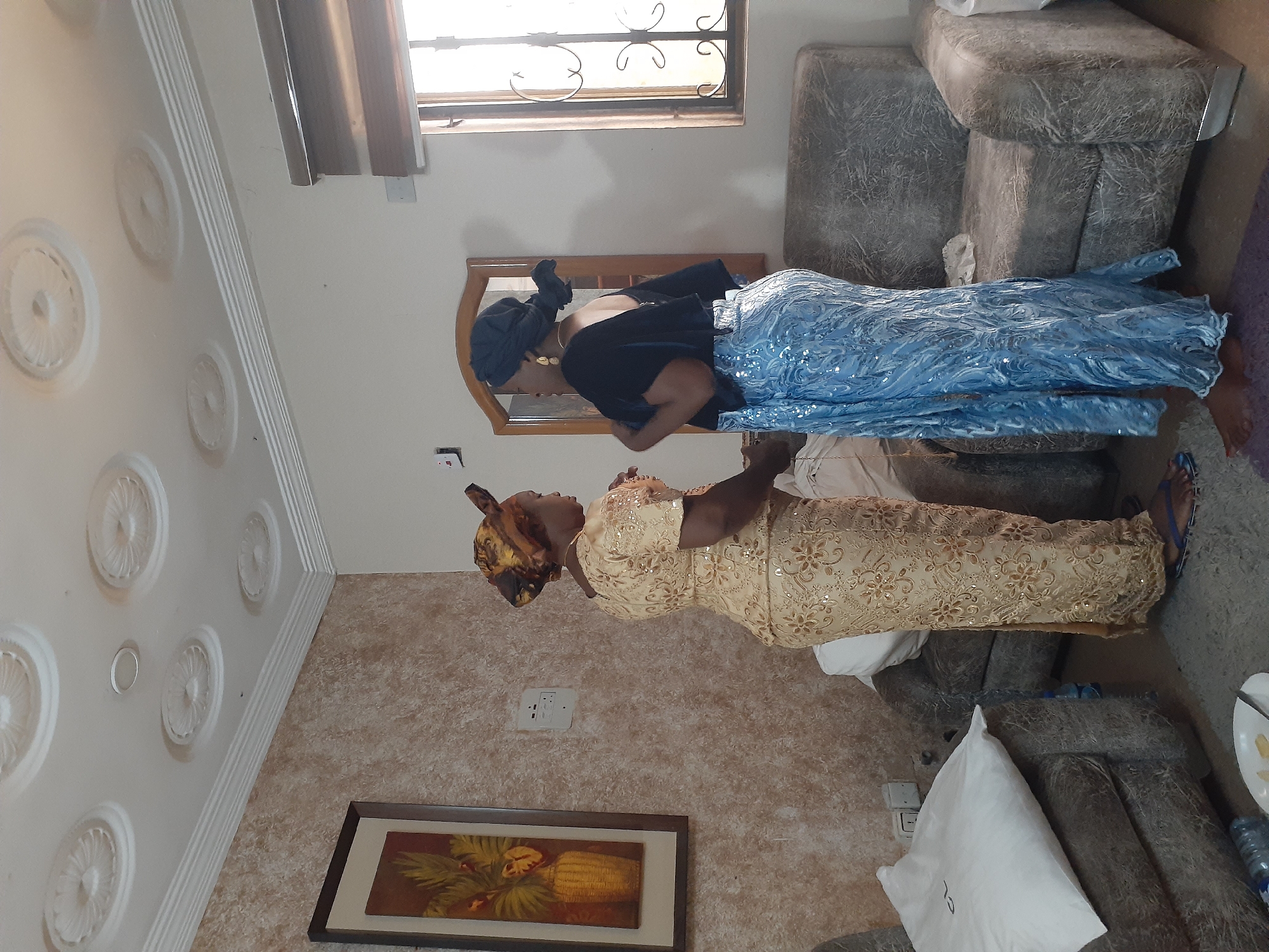 In a brightly lit interior in Jos, Nigeria, two women are the central subjects. One woman, dressed in a blue sequined dress and a black top, stands by a mirror, appearing to look at her reflection. The second woman, wearing a golden dress, stands nearby, facing the first woman. A beige sofa is positioned in the background. The room also features ornate ceiling detailing, a framed picture on the wall, and a window with metal bars.