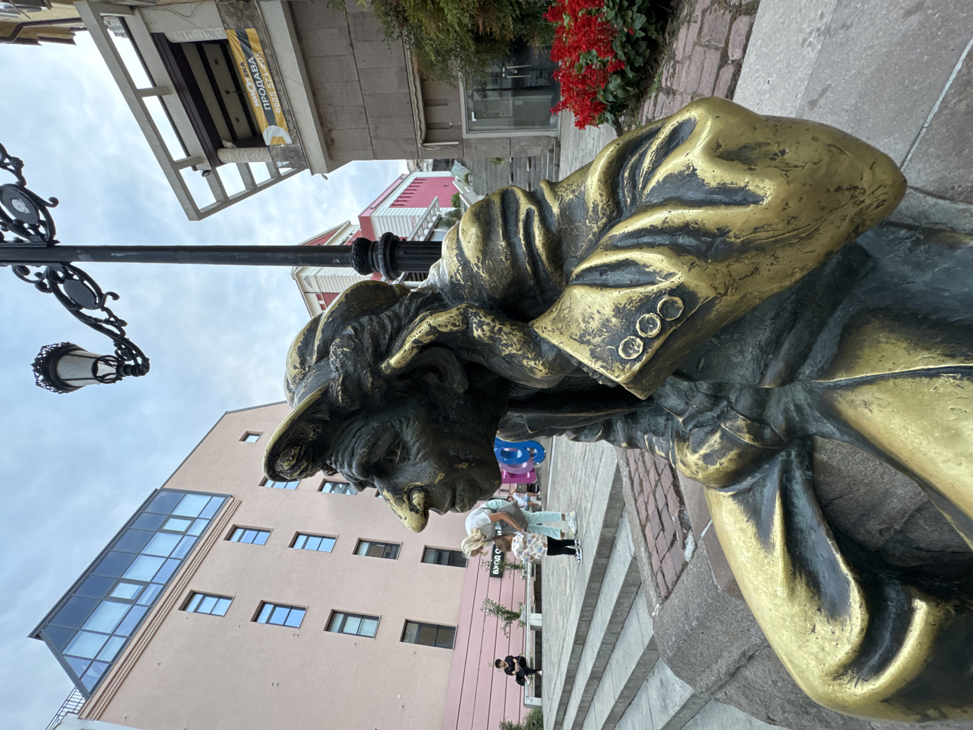 A bronze statue of a man wearing a cap and jacket is the primary subject. He is depicted in a seated pose, with his right hand touching his ear. The statue has a weathered, aged appearance with a golden patina. In the background, a light pink building with modern glass windows and a mansard roof is visible. To the right of the statue, a building with a balcony displays a banner advertising real estate for sale. A lamppost with an ornate design stands behind the statue. In the mid-ground, several people, including a woman with a child, are interacting on a set of stone steps. A bench with a person sitting on it is also present. The ground is paved with cobblestones. The scene appears to be an urban setting. The statue is located on the pedestrian bridge connecting the central part of Plovdiv with the Tsar Simeon's Garden.