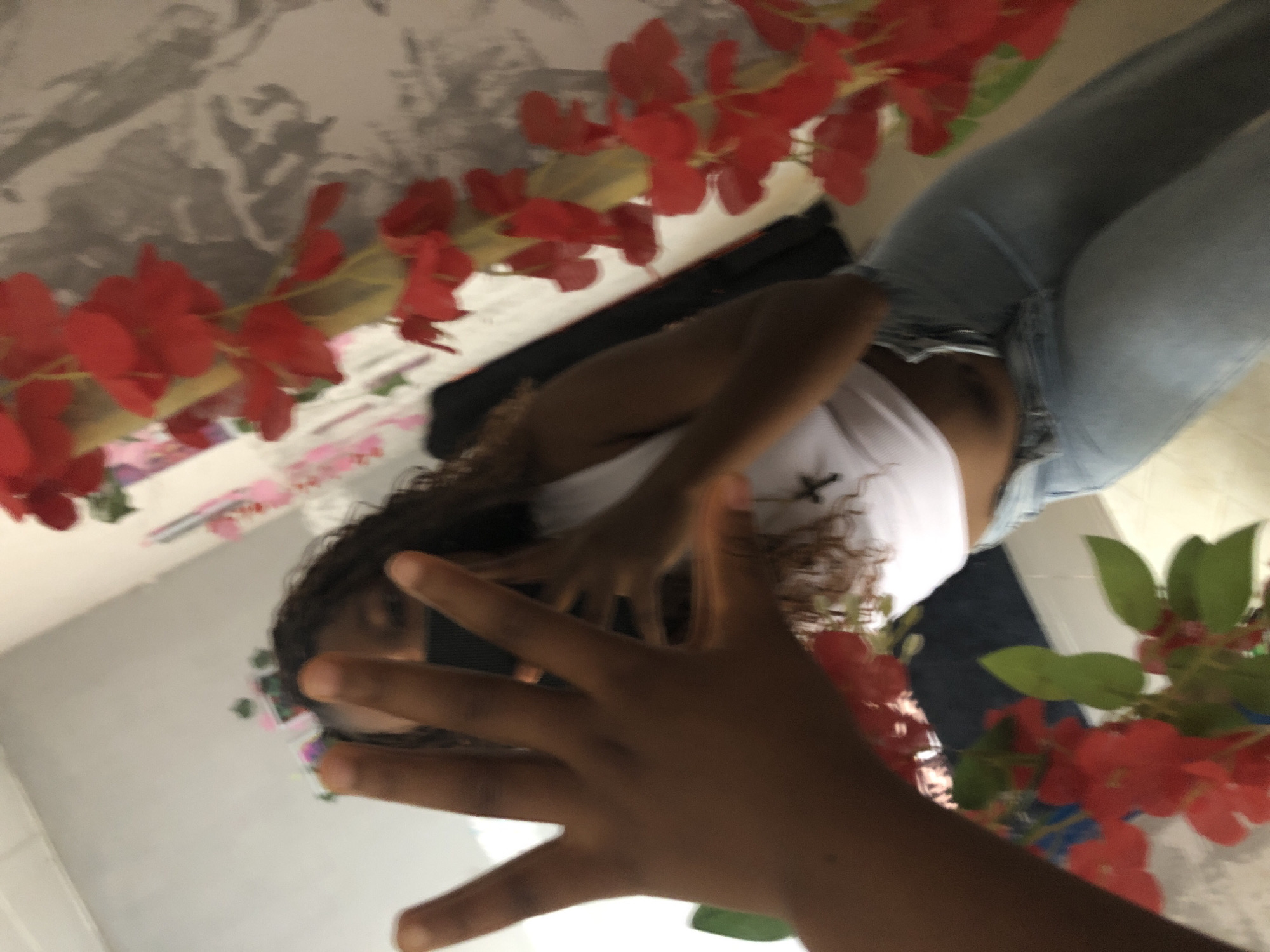 An overhead shot, possibly a mirror selfie, shows a dark-skinned person with curly hair wearing a white tank top and light blue jeans. The person’s hand is prominently in the foreground. Red flowers surround the subject. The background includes a wall with an indistinguishable pattern or picture on it. The location in Kashere, Nigeria, cannot be verified from the image.