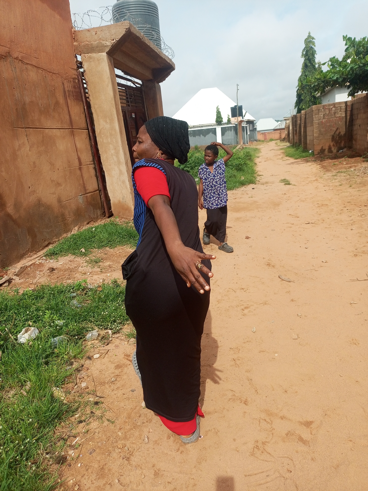 A woman wearing a black headscarf, red shirt, and a long black dress with a blue stripe, faces away from the camera and towards the right. She is standing on a dirt path. Her left arm is outstretched, and her left hand, adorned with rings, is visible. Beside her, to the left, is a dirt wall. In the background, a young girl in a blue and white patterned shirt and black pants stands with her hand on her head. Behind the girl, a street lined with brick walls and trees extends into the distance under a cloudy sky. The scene appears to be an outdoor environment within the city of Tempure, Nigeria.
