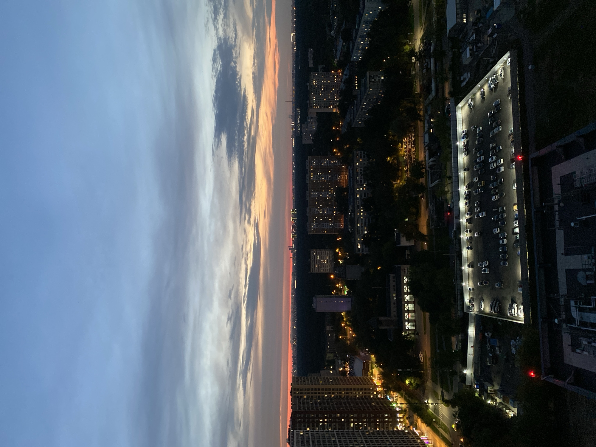 This is an aerial, low-light, wide-angle photograph taken from a high vantage point, looking out over a cityscape at dusk or dawn. The sky exhibits a gradient of colors, transitioning from a deep blue at the top to lighter blues and soft oranges and pinks near the horizon, with scattered, wispy clouds.
Dominating the mid-ground and foreground is a dense urban environment with numerous apartment buildings of varying heights. Many windows in these buildings are illuminated, suggesting the presence of residents. One prominent, tall, multi-story residential building is visible on the left side of the image, its facade brightly lit by streetlights and interior lights.
In the foreground, a large, well-lit parking lot is visible, filled with numerous parked cars. The parking lot appears to be enclosed by a fence, and several lamps illuminate the area. To the left of the parking lot, a street with some traffic and sidewalks can be seen, also illuminated by streetlights. Further into the background, a dark expanse of trees suggests a large park or forest area separating different parts of the city. Beyond the trees, the distant city skyline is dotted with lights and the silhouettes of taller structures, including what appear to be industrial or power plant facilities.
The overall scene depicts a typical residential and urban environment during twilight. Given the architectural styles and the density of the apartment buildings, this location is likely a residential district in Moscow, Russia. The presence of numerous illuminated windows and parked cars indicates a populated area. There are no visible people or specific actions being performed. The environment is characterized by artificial lighting from buildings and streetlights contrasting with the natural light from the sky.