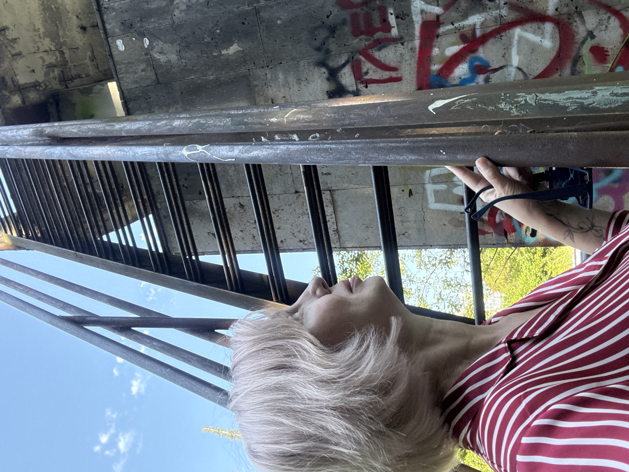 The image, oriented correctly, depicts a person with short, light-blonde hair looking upwards, situated outdoors beneath a large concrete structure, likely a bridge or overpass. The individual is holding onto a metal railing with their right hand, and a pair of black sunglasses hangs from their wrist. They are wearing a red and white striped collared shirt.
Above the person, the underside of the concrete structure is visible, adorned with extensive stylized red and black graffiti. Some of the markings appear to be abstract or unidentifiable letters and tags. The metal railings of a staircase or platform extend vertically through the frame. In the background, a clear blue sky with a few faint white clouds is visible, along with glimpses of green foliage, suggesting a bright, sunny daytime setting. The concrete surfaces show signs of weathering and discoloration. The scene provides an impression of an urban or semi-urban environment in Sochi, Russia.