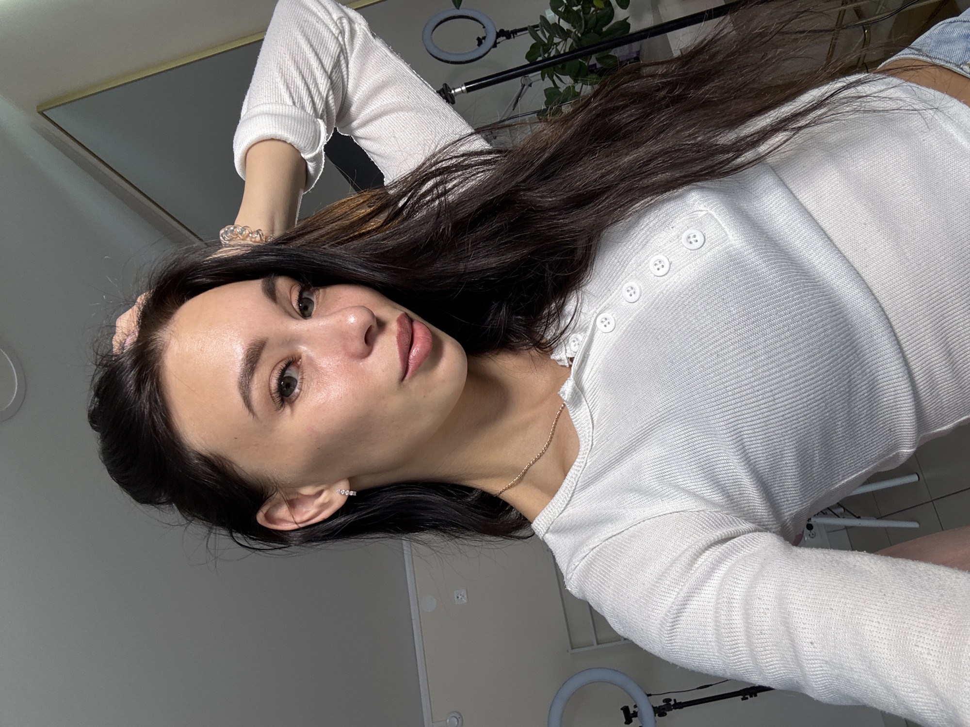 A young woman is posing for a selfie in a room with a ring light and some indoor plants visible in the background. She is wearing a white long-sleeved, button-up shirt, jewelry on her wrist and neck, and a small earring in her left ear. The room has a mirror on the wall. Based on the presence of what appears to be a Russian-style power outlet on the wall and previous requests, the location can be assumed to be Chaikovsky, Russia.