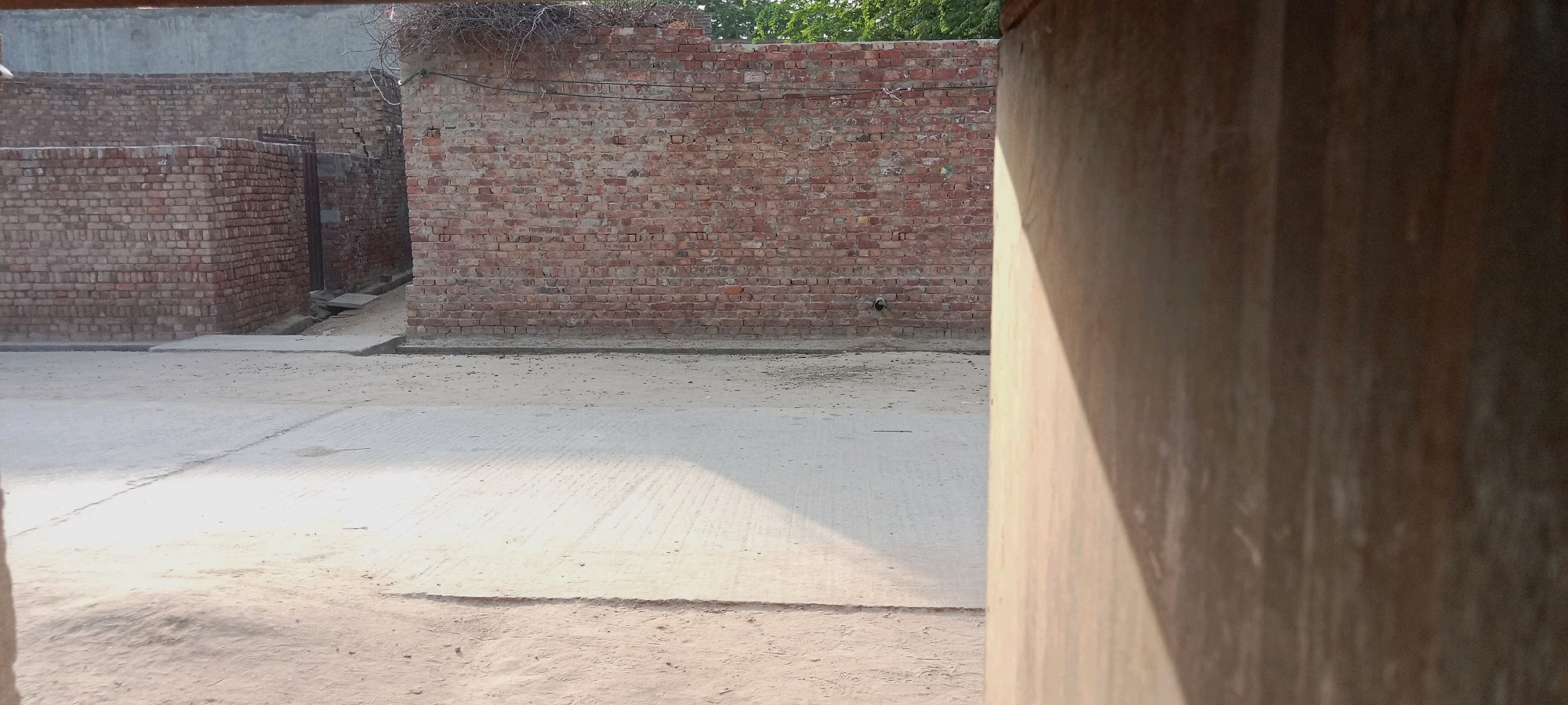 A dusty, unpaved road runs through the foreground, with a concrete path intersecting it from left to right. The road is flanked by brick walls, suggesting an urban or semi-urban environment in Jatri Kohna, Pakistan. In the mid-ground, a large, weathered brick wall dominates the scene. The wall has a rough, uneven texture, with visible mortar lines between the bricks. A small, dark object, possibly a drainpipe or a vent, is embedded in the wall. A narrow, unpaved alleyway is visible to the left of the main wall, leading into a darker recess. The upper portion of the image reveals a sliver of green foliage, indicating the presence of trees or vegetation beyond the structures. The right side of the frame is occupied by a large, vertical wooden beam or pillar, casting a distinct shadow across the scene. The overall impression is one of an old, possibly rural or informal settlement, characterized by its rudimentary construction and arid surroundings. No people or animals are visible in the frame.