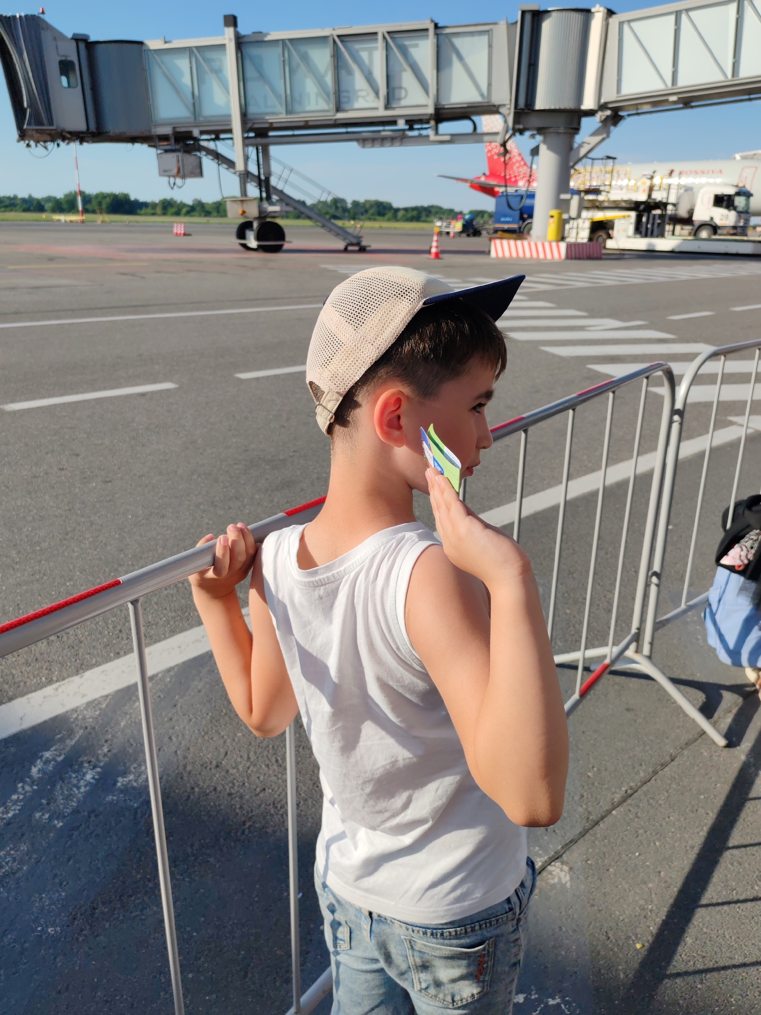 A young boy, wearing a white t-shirt and jeans, stands at an airport, holding onto a metal railing. He has a beige baseball cap on his head. The boy is looking to his right, and has several colorful paper airplanes stuck to his ear. In the background, a jet bridge extends from an airport terminal to a parked airplane. The sky is clear and blue, suggesting it is daytime. The scene appears to be at an airport, likely in Khrabrovo, Russia, given the provided location context.