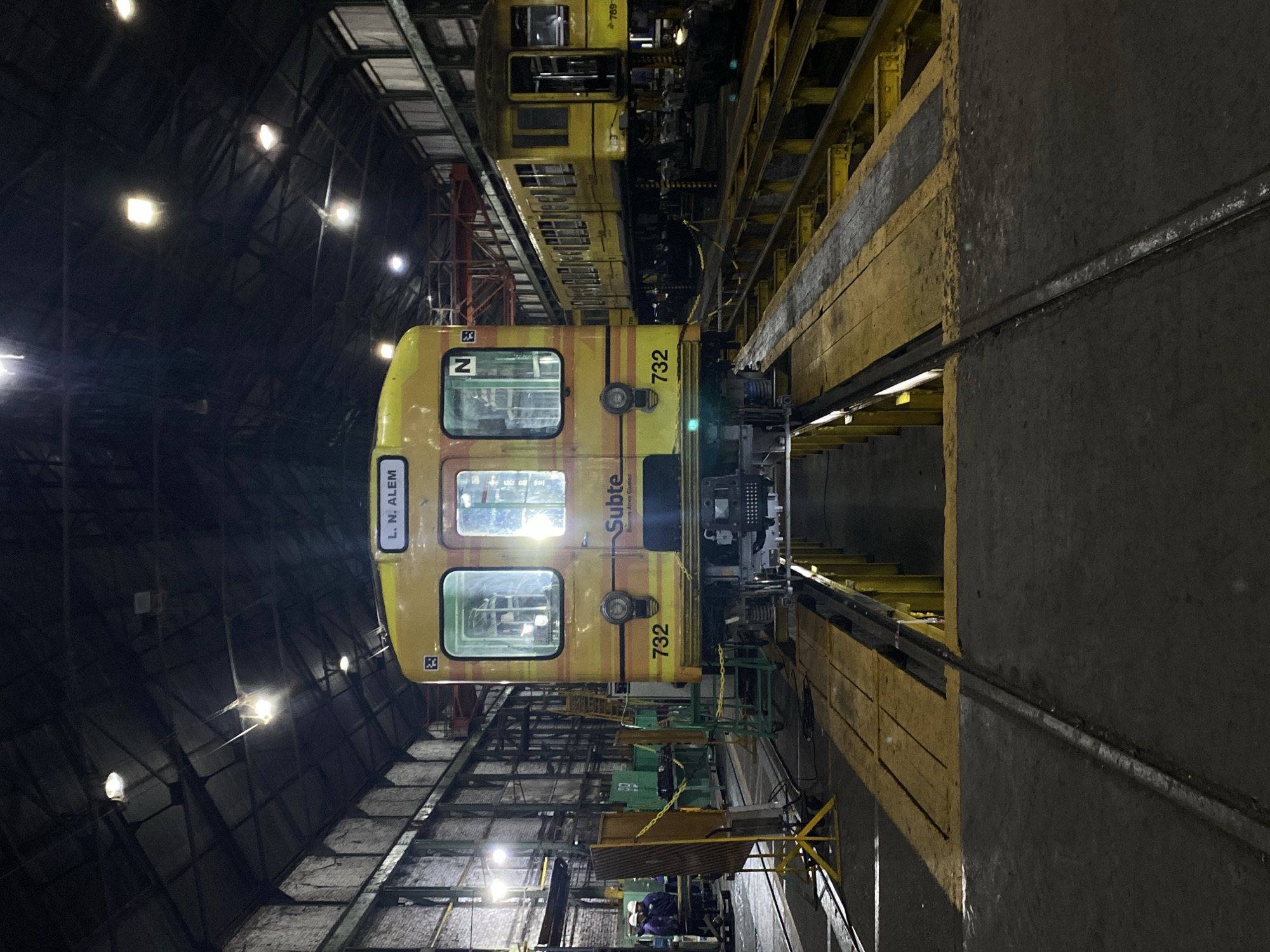 The image depicts a yellow Subte (subway) train inside a maintenance depot in Buenos Aires, Argentina. The train car, numbered 732, is labeled "Subte" and has "L. N. Alem" visible on its side, indicating a destination. The depot has a high ceiling with multiple lights illuminating the scene. The tracks are visible below the train, and maintenance equipment and personnel wearing hard hats are present in the background. The environment suggests an industrial setting focused on the upkeep of subway cars.