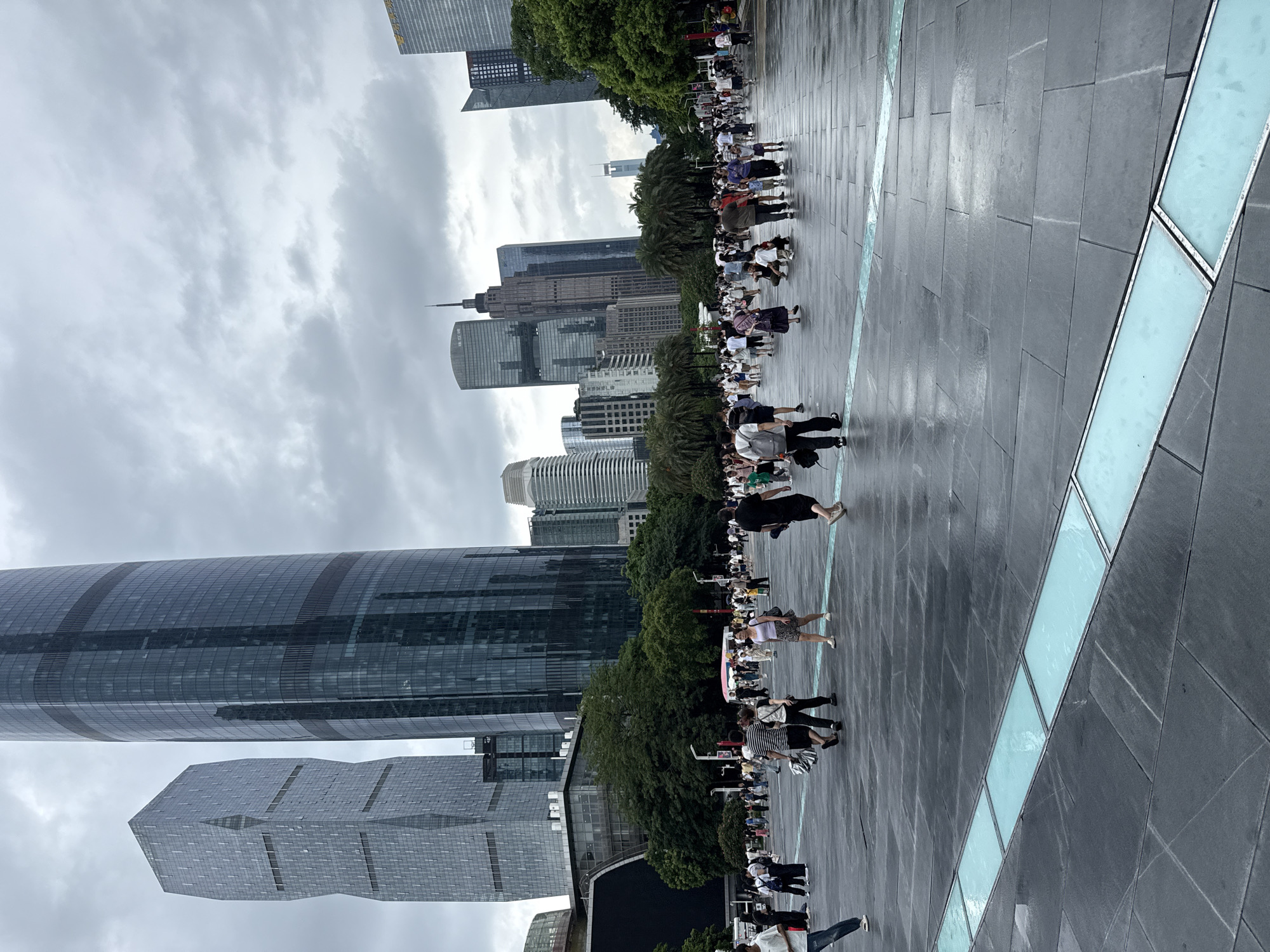 The image depicts a wide, paved plaza in Tianhe District, China, on a cloudy day. Several modern skyscrapers with glass facades dominate the background, with the distinctive rounded International Finance Centre and the angular CTF Finance Centre prominently visible. A crowd of people are walking and standing in the plaza, some carrying bags or pushing strollers. The ground is wet and reflective, suggesting recent rain. A section of the plaza features inlaid rectangular panels with a light blue, translucent material. The overall scene is one of urban activity and modern architecture under an overcast sky.
