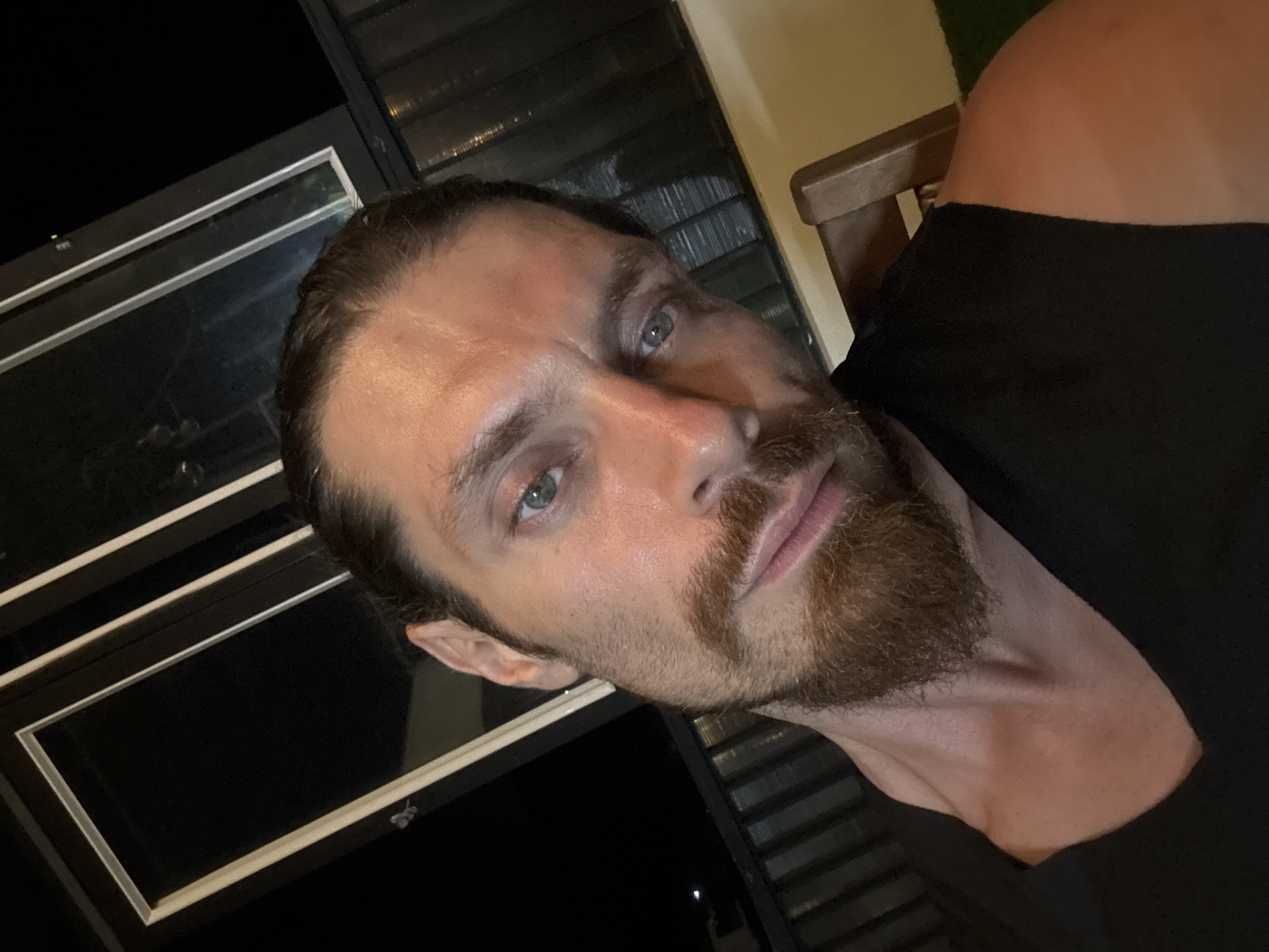 A close-up, eye-level, selfie-style shot shows a man with medium-length, dark brown hair pulled back and a neatly trimmed beard. He is wearing a sleeveless black shirt. His skin appears to have a healthy glow. He has light blue eyes and is looking directly at the camera with a slight, subtle smile.
The man is positioned in the foreground, with a dimly lit background suggesting an outdoor evening setting. Behind him, a dark railing is visible, partially obscured by his right shoulder and the right edge of the frame. Further back, a wooden chair is visible on the right side of the image. Above the railing, a window with dark panes and white trim is prominent, reflecting some light. The overall lighting suggests ambient light sources, possibly from the window and other unseen sources, creating a moody atmosphere. The scene is captured at night.