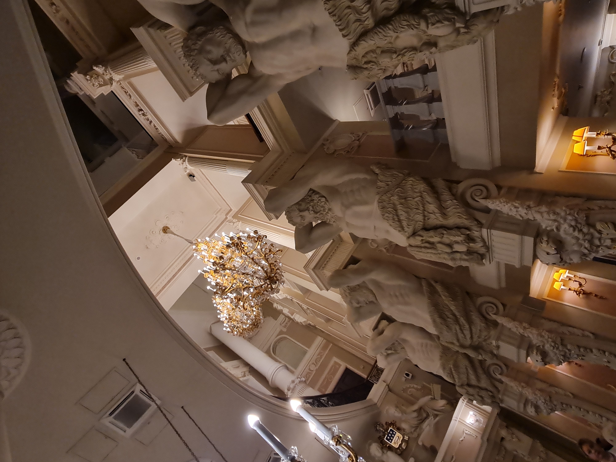 The image depicts the interior of the Grand Duke Vladimir Alexander Palace in Saint Petersburg, Russia. The scene features ornate architecture with classical columns and decorative elements. Prominently displayed are several large, white statues of muscular male figures with goat-like legs, supporting architectural elements above them. These figures appear to be caryatids or telamones, rendered in a classical style. Above the statues, a large, elaborate crystal chandelier hangs from the ceiling, casting a warm light. In the background, more architectural details, including arches and further columns, are visible, along with a glimpse of a balcony railing. The lighting is dim, accentuating the grandeur of the decor and the statues. There are no visible actions or interactions taking place.
