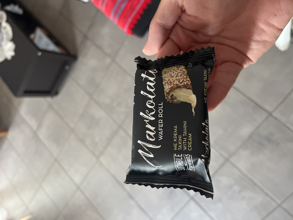 A hand is holding a package of Markolatu Wafel Roll with tahini cream. The package is black with white writing. The writing on the package says "Markolatu Wafel Roll, Me Krema Taxini with Tahini Cream." The hand is holding the package in front of a white tiled floor. The coordinates (40.627, 24.575) correspond to a location in Limenaria, Greece. This location is likely an indoor space, potentially a residential home. The environment is typical of a residential setting, with a tiled floor and a hand holding a food product. The image was likely captured by a person to document the product.
