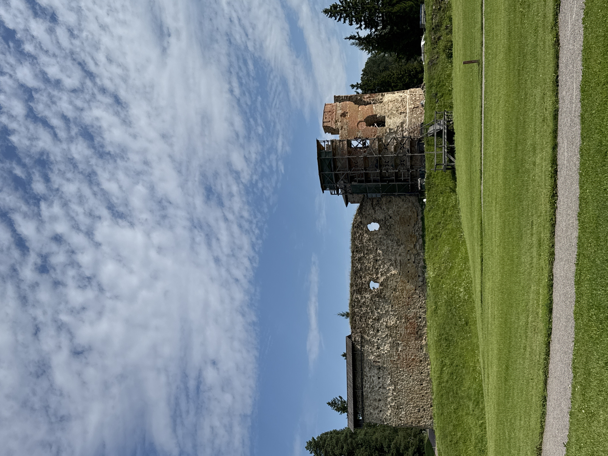 This image captures the ruins of Vana-Vastseliina Castle in Vana-Vastseliina, Estonia. The ancient stone and brick structure, showing signs of decay and ongoing restoration with scaffolding on one of the towers, stands against a bright blue sky dotted with white, wispy clouds. A lush green lawn slopes up towards the castle walls, with a gravel path winding through the foreground. Tall evergreen trees are visible behind the ruins, suggesting a natural, park-like setting. The scene is peaceful and bathed in sunlight, indicating a clear day.