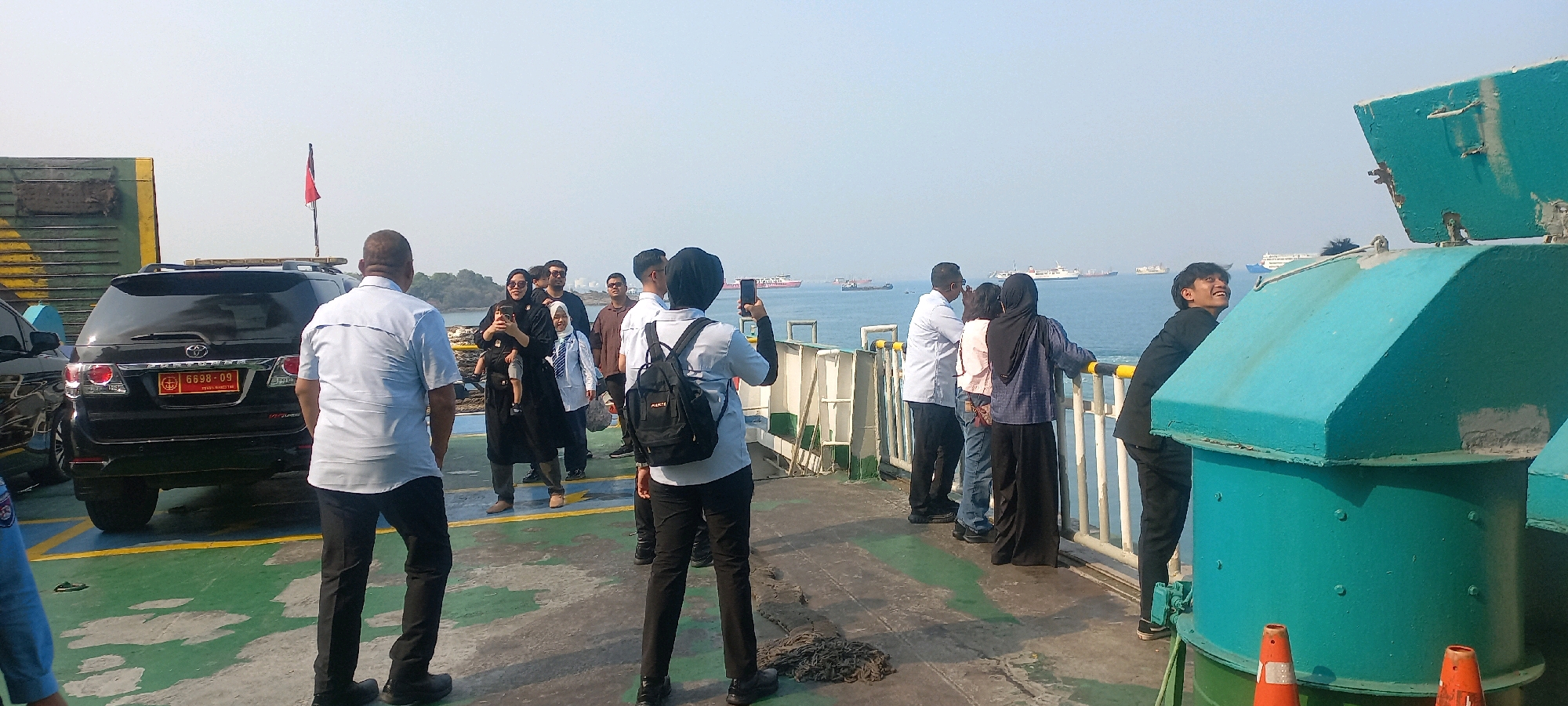 The image depicts a group of people on a ferry in Cilegon, Indonesia, with the city's coastal landscape visible in the background. A black SUV with a license plate reading "6698-09" is parked on the ferry deck. Several individuals are standing near the railing, observing the sea and other vessels, while some are taking pictures with their phones. One person is holding a baby. The sky is clear and the water appears calm. The ferry deck is marked with yellow lines and a green surface. The overall scene suggests a journey or excursion, with people enjoying the view.