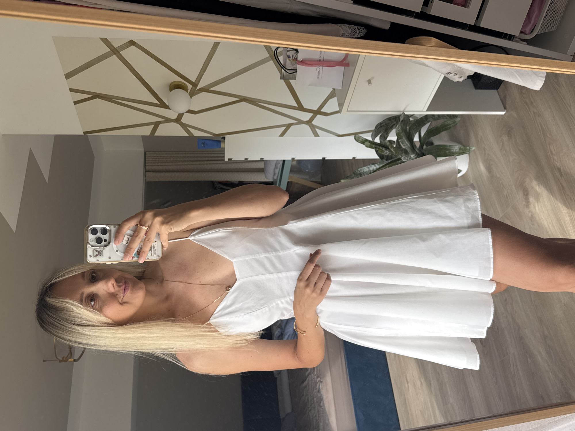 A woman with blonde hair is taking a mirror selfie in what appears to be a bedroom. She is wearing a white spaghetti strap dress and gold jewelry, including a necklace and a bracelet. The room has a modern design with a geometric wallpaper and a tall mirror. In the reflection, a bed with blue bedding is visible, along with a dresser and a houseplant. The overall lighting suggests it is daytime.
