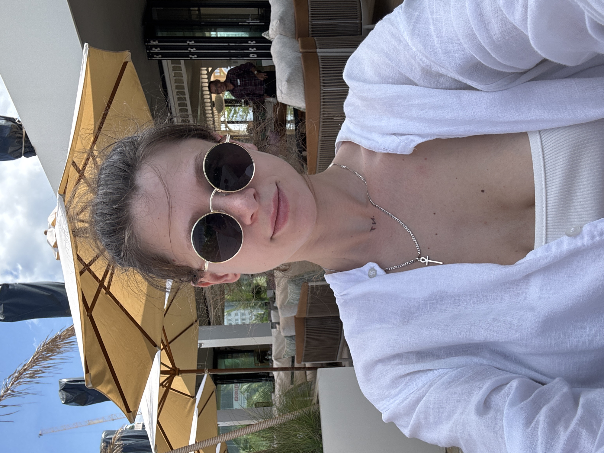 The image shows a woman wearing round sunglasses and a white shirt, likely linen, standing outdoors, likely in Đà Nẵng, Vietnam. In the background, there's an outdoor seating area with parasols and lounge furniture, possibly a hotel or resort. Another person is visible further in the background, seemingly walking. A building is visible behind the seating area. The sky is visible in the background with a construction crane. The woman appears to be taking a selfie. She is wearing a silver necklace with a pendant. A tattoo is visible on her neck.