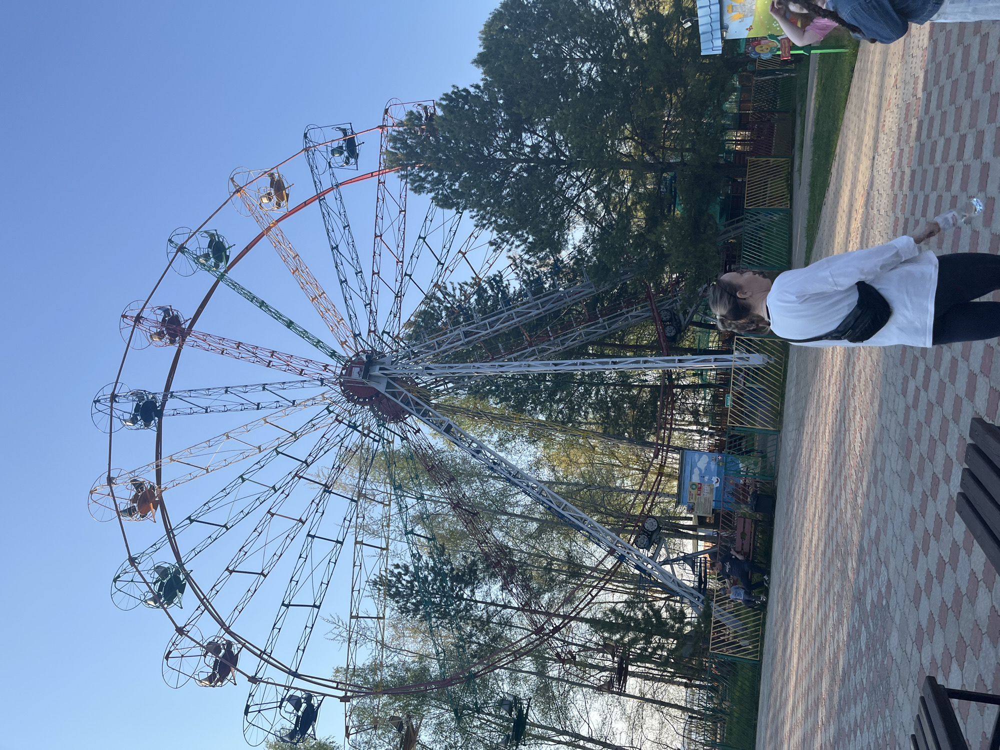 The image shows a Ferris wheel in a park in Chaikovsky, Russia. The Ferris wheel is tall and brightly colored with rainbow hues on the spokes and gondolas. People are riding in some of the gondolas. The sky is clear and blue. Tall trees stand behind the Ferris wheel. In the foreground, a woman in a white shirt and black pants stands on a brick-patterned walkway, looking up at the Ferris wheel. The walkway has a checkerboard pattern of red and beige bricks. A bench sits next to the walkway. Part of another person is visible near the top of the image. A fence surrounds the park area. The photo is taken in daylight, giving the scene a clear, crisp appearance.