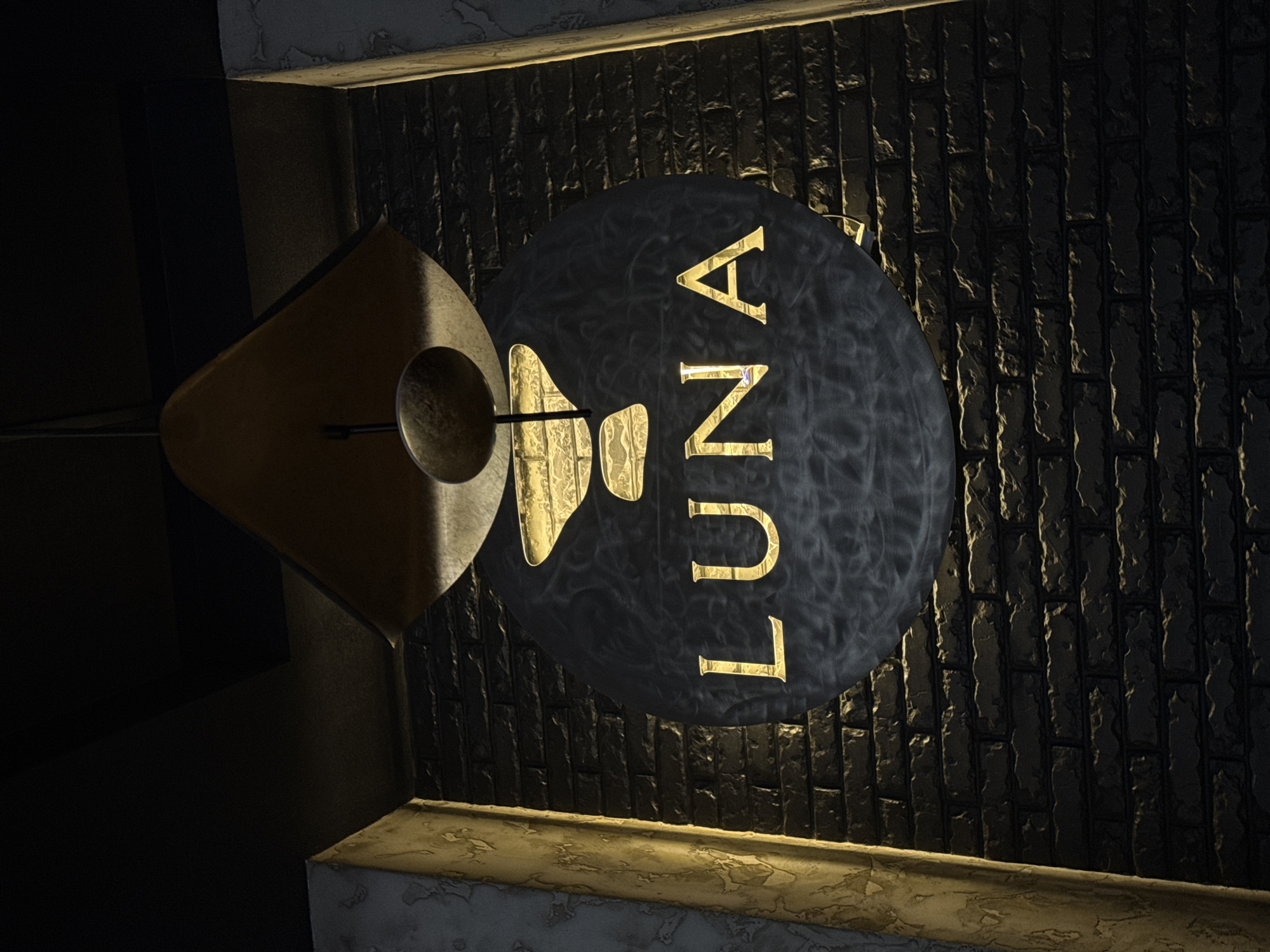 The image shows a sign for a business named "LUNA." The sign is circular, dark in color with the name vertically oriented and in golden letters. The background appears to be a textured brick wall. A geometric light fixture is seen to the left of the sign. The location is Unknown, Unknown.