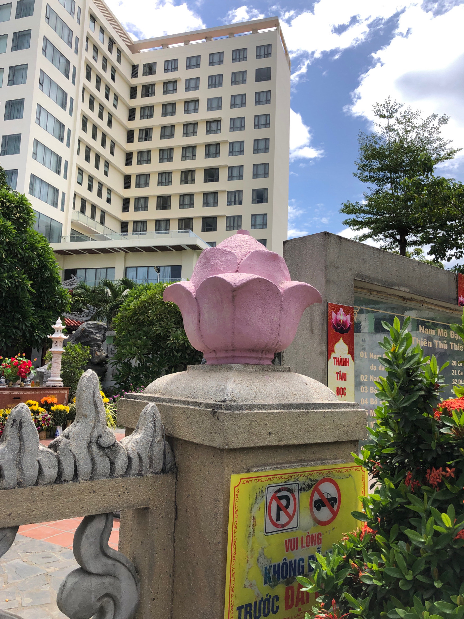The image depicts a scene in Đà Nẵng, Vietnam, featuring a modern multi-story hotel building in the background with a cloudy blue sky above. In the foreground, there is a pink lotus-shaped statue atop a stone pedestal. To the right of the statue, a concrete wall has a sign with Vietnamese text and lotus motifs, suggesting it may be part of a temple or spiritual site. Below the lotus statue, a yellow sign with red and white symbols prohibits parking and vehicles. Green foliage and red flowers are visible around the base of the concrete wall and sign, and a decorative grey stone fence is in the lower left of the image. The overall impression is a blend of modern architecture, traditional elements, and natural greenery.