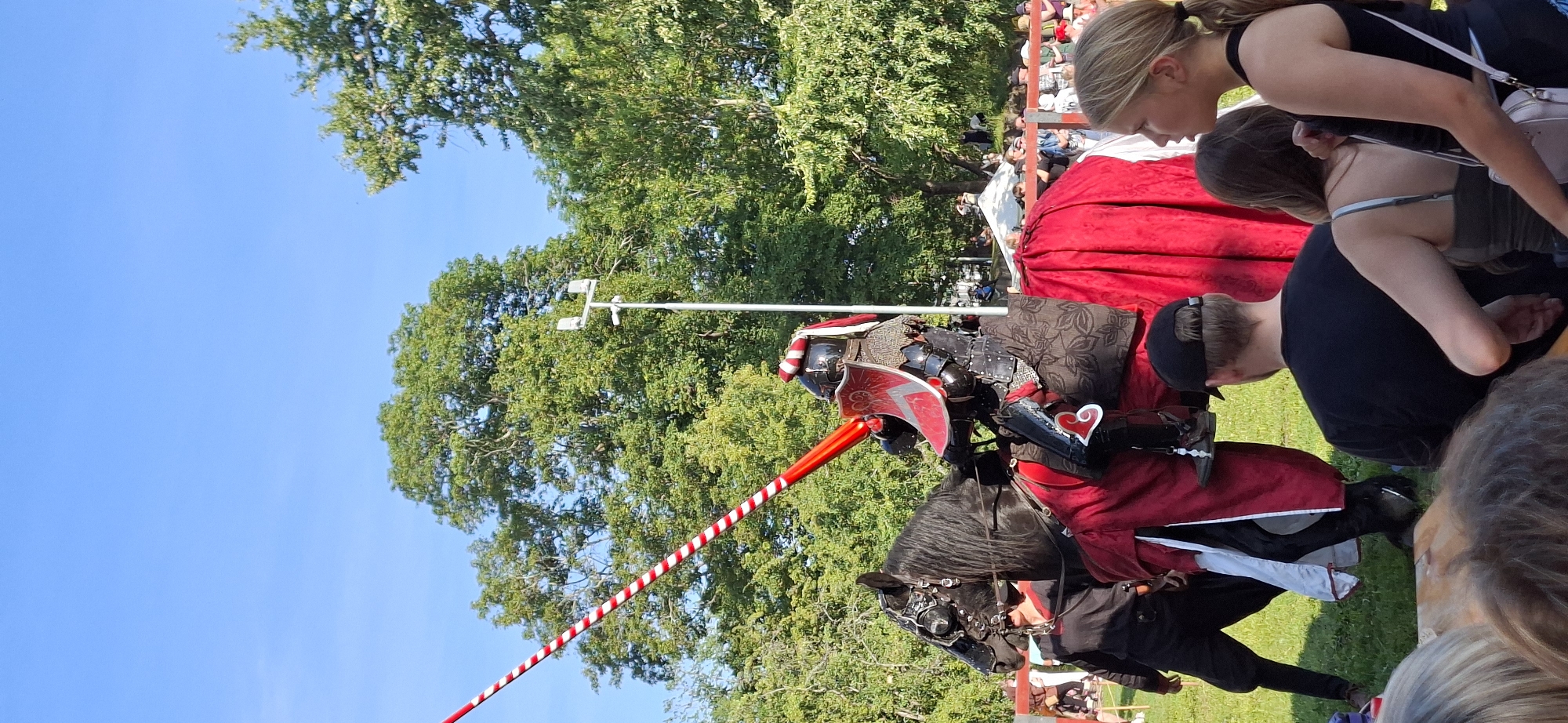 This image captures a jousting reenactment event, likely at the Turku Medieval Market, given the historical theme. In the foreground, spectators are visible, their backs turned to the camera, suggesting they are facing the jousting arena. A young woman with blonde hair tied in a ponytail is looking to the side.
The central subject is a knight in full black armor, mounted on a black horse. The knight holds a long lance with red and white striped detailing, pointed towards the viewer's left. The horse is adorned with a red caparison, and the knight's armor includes a red and silver shield. Another person, possibly a squire, dressed in black attire, stands to the left of the horse, holding the reins.
The background consists of lush green trees under a clear blue sky, typical of an outdoor event in a park or open field. There are indications of other attendees and stalls further in the background, subtly suggesting a larger festival atmosphere. The presence of a light pole with lighting fixtures in the middle ground indicates a modern event setup within a potentially historical reenactment.